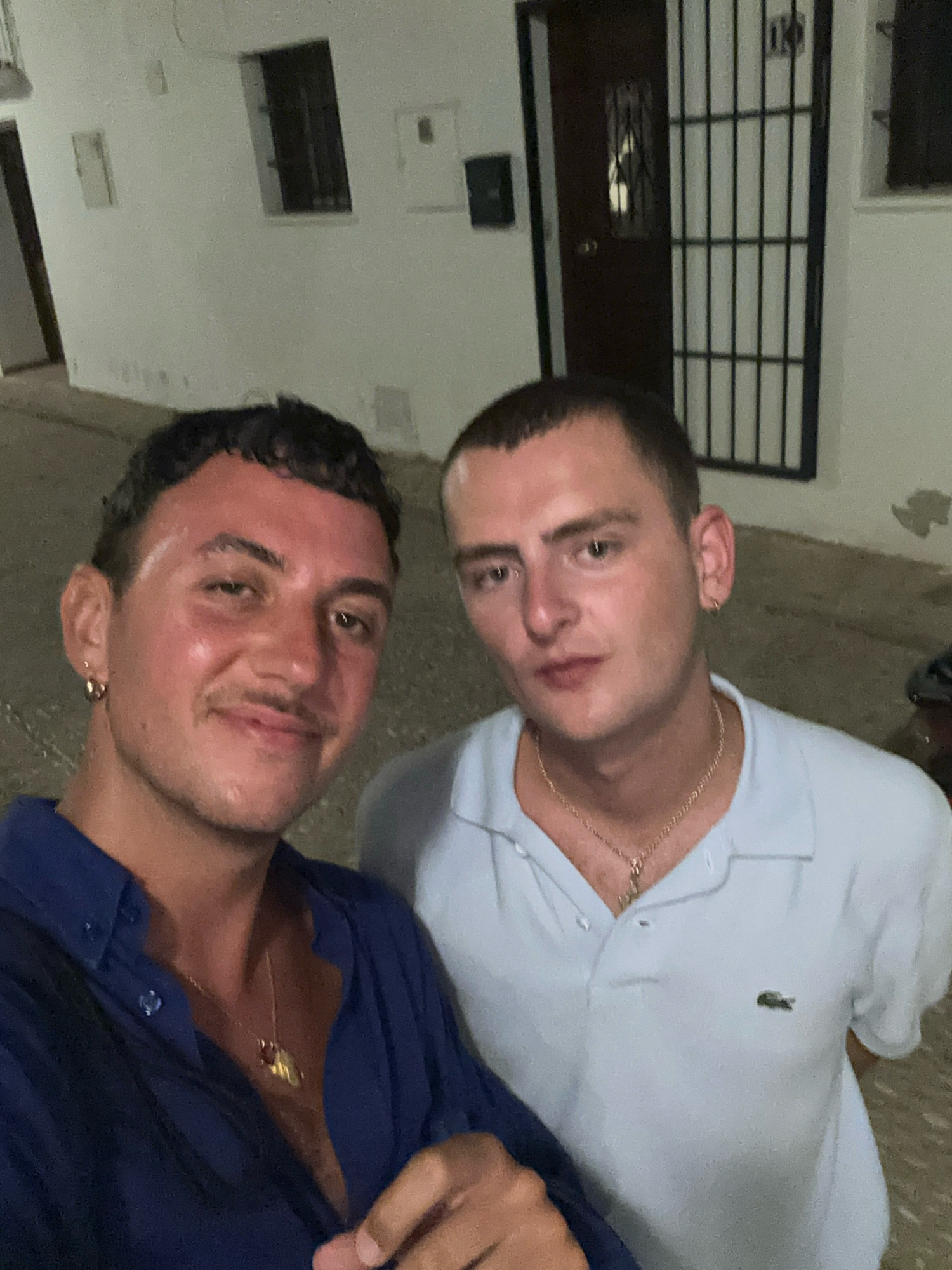 Two young men are posing for a selfie outdoors at night, in the city of Altea, Spain. The man on the left is wearing a dark blue open-collared shirt and a gold necklace. He has short, dark curly hair, a mustache, and a gold hoop earring in his left ear. The man on the right is wearing a light blue polo shirt with a small green crocodile logo on the left side of his chest and a gold necklace. He has short, light brown hair and a small gold earring in his right ear. The background shows a white building with a dark brown door protected by a metal gate, and a narrow street. The overall lighting suggests it is evening or night.