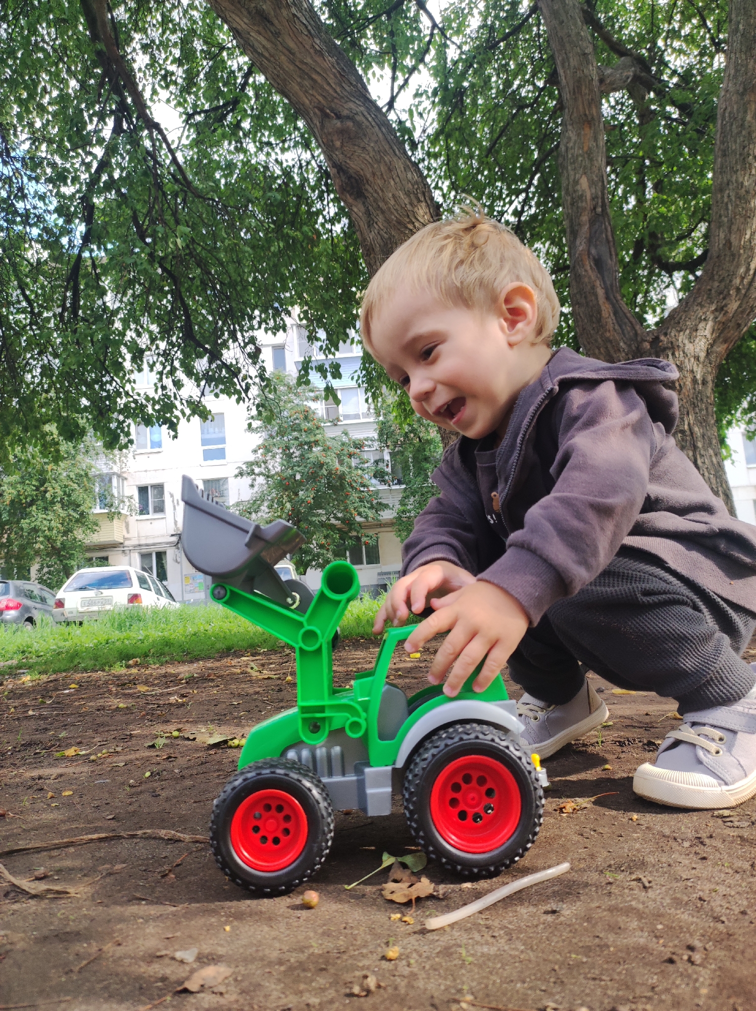 A young Caucasian boy with blonde hair, wearing a dark hooded jacket and dark pants, kneels on the ground and plays with a green and gray toy tractor. The tractor has red-rimmed wheels. The boy is looking down at the toy with a happy expression. The scene takes place outdoors in an urban environment, with a large tree providing shade. In the background, there are apartment buildings and cars. The ground is covered with dirt and fallen leaves. The image appears to be taken on a sunny day.