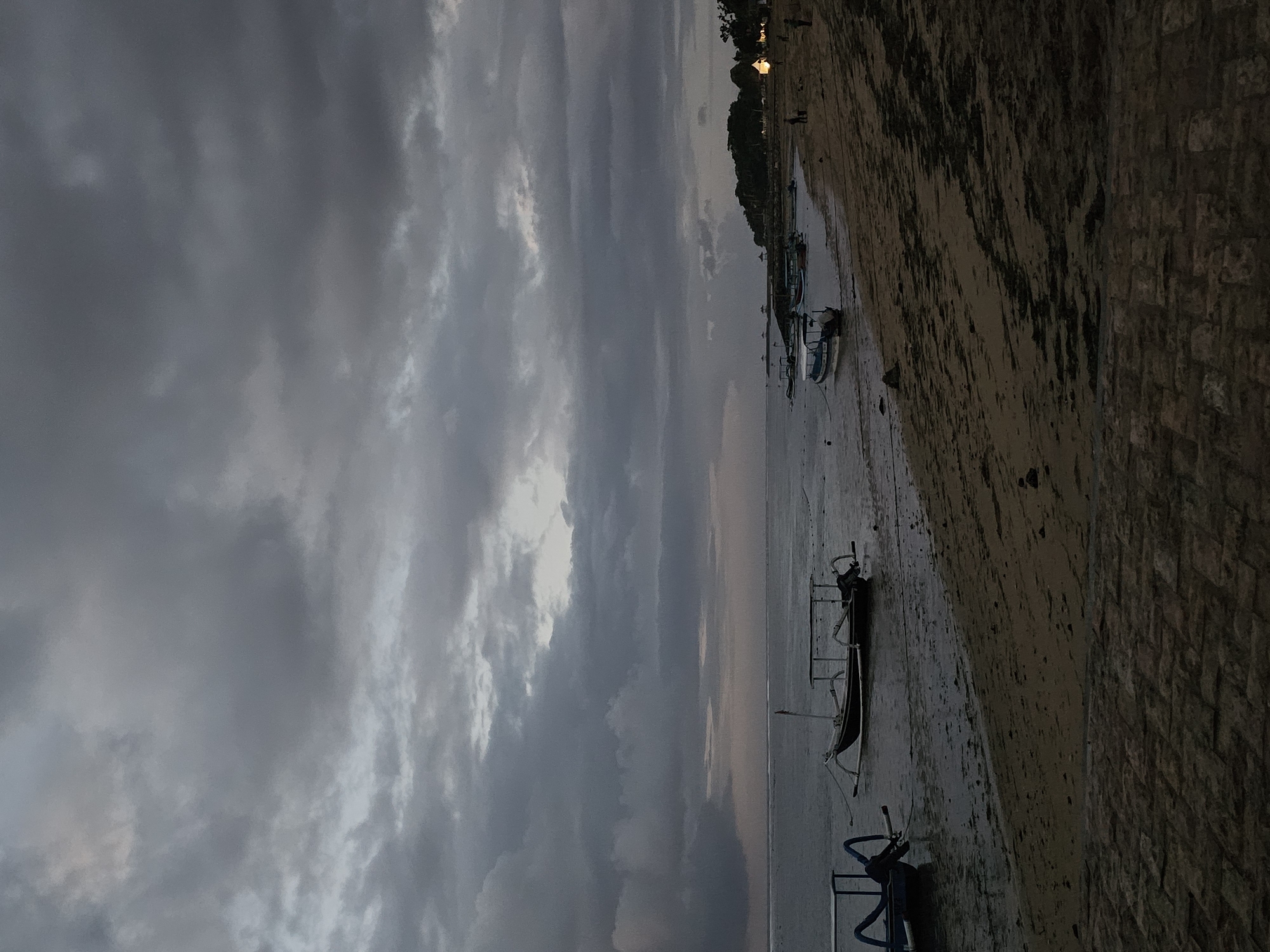 This image depicts a coastal scene at dusk, likely in Denpasar, Indonesia. The sky is overcast with heavy, grey clouds that dominate the upper portion of the frame, casting a somber mood over the landscape. The sea is calm, with gentle waves lapping at the sandy shore.
Several traditional fishing boats are anchored in the shallow water near the beach. These boats, characterized by their outriggers and simple designs, are a common sight in Indonesian coastal villages. The beach itself is sandy, with patches of darker seaweed or debris scattered across the wet sand near the water's edge.
In the distance, along the coastline, there is a line of trees and vegetation, suggesting a developed area. A few small lights are visible among the trees, indicating human habitation or businesses. A paved pathway or walkway made of stone is visible in the foreground, leading towards the beach. The overall atmosphere is tranquil, suggesting the end of a day or the start of a quiet evening.
