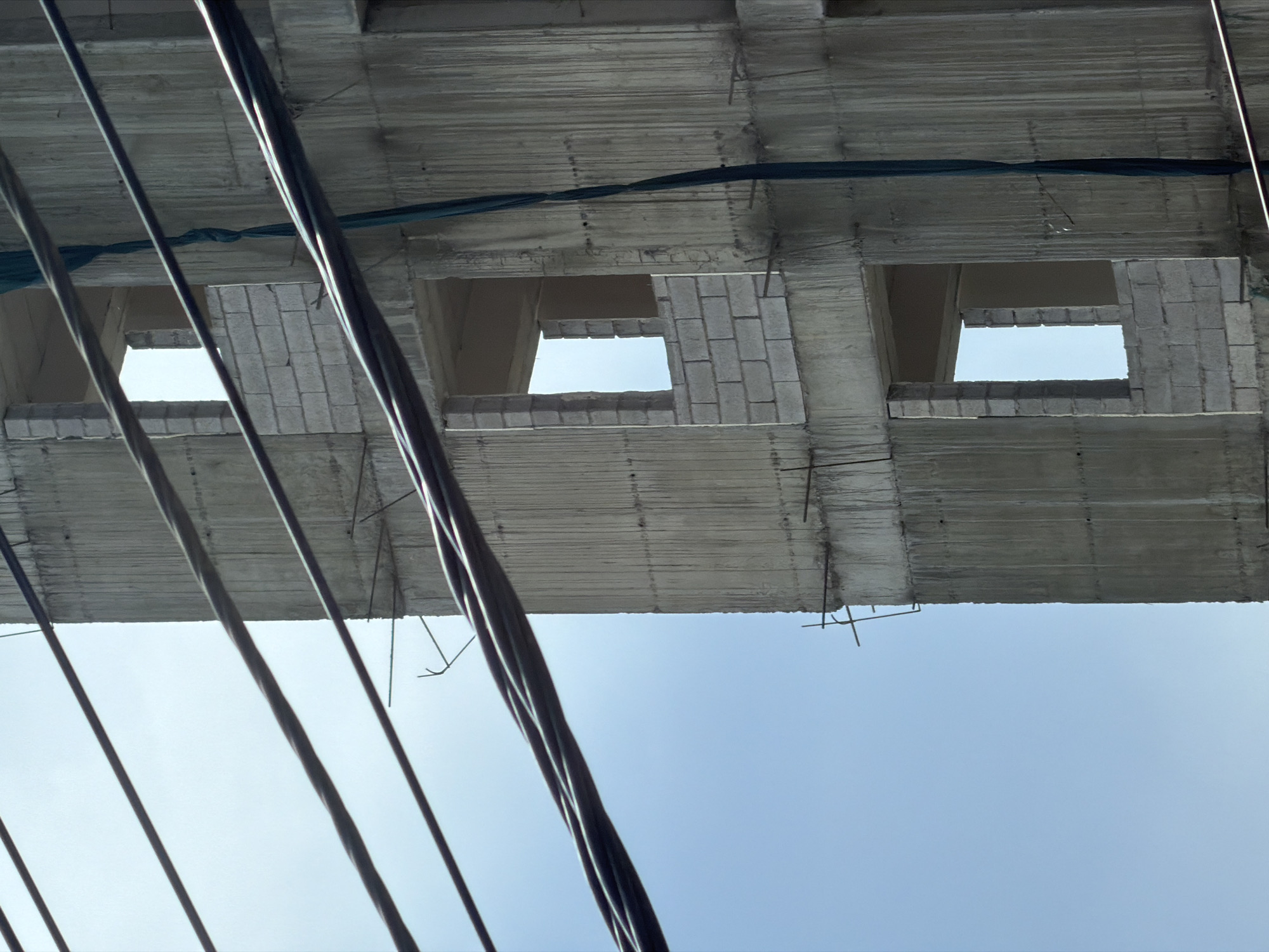 The image captures an upward view of an unfinished concrete structure, likely part of a building under construction, set against a clear, light blue sky. The scene is located in Yerevan, Armenia.
The primary subjects are the raw concrete underside of a floor or elevated platform, and multiple dark, thick cables or wires running diagonally across the lower and middle foreground. The concrete surface displays texture from its formwork and has visible rebar (reinforcing steel bars) protruding in various places, indicating the incomplete state of construction. Several square or rectangular openings are visible in the concrete structure, possibly designated for windows or architectural features. One such opening features light-colored bricks framing a section of its interior. A piece of dark blue or green fabric, possibly netting, is draped and tied around a beam section in the upper part of the concrete structure.
No people or direct construction activity are visible, but the raw materials and exposed rebar strongly suggest ongoing building work. The clear blue sky indicates it is daytime with good weather conditions. The perspective is from directly below the structure, giving an unusual and abstract view of the construction. No text is visible in the image.