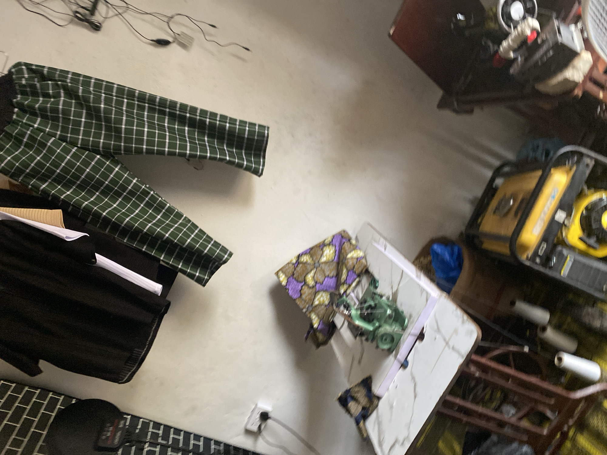 The image depicts an indoor scene, likely a workshop or a domestic space, in Jalingo, Nigeria. In the upper portion of the frame, a black t-shirt is hanging, alongside a pair of green checkered trousers. To the left, a black cap with white lettering is visible. A white electrical outlet with a plug is mounted on the wall.
In the lower half of the image, a sewing machine, appearing to be green, is positioned on a white table with a marble-like pattern. To the right of the sewing machine, a colorful piece of fabric, patterned with purple and gold floral designs, is folded. A small, dark patterned fabric piece is to the left of the sewing machine.
Below the table, there are several white cylindrical spools, possibly of thread or a similar material. A yellow generator is located to the right of the table. Further to the right, a dark red chair or stool is partially visible. The overall environment appears somewhat cluttered, suggesting a functional workspace. The lighting is dim, casting shadows on the walls.