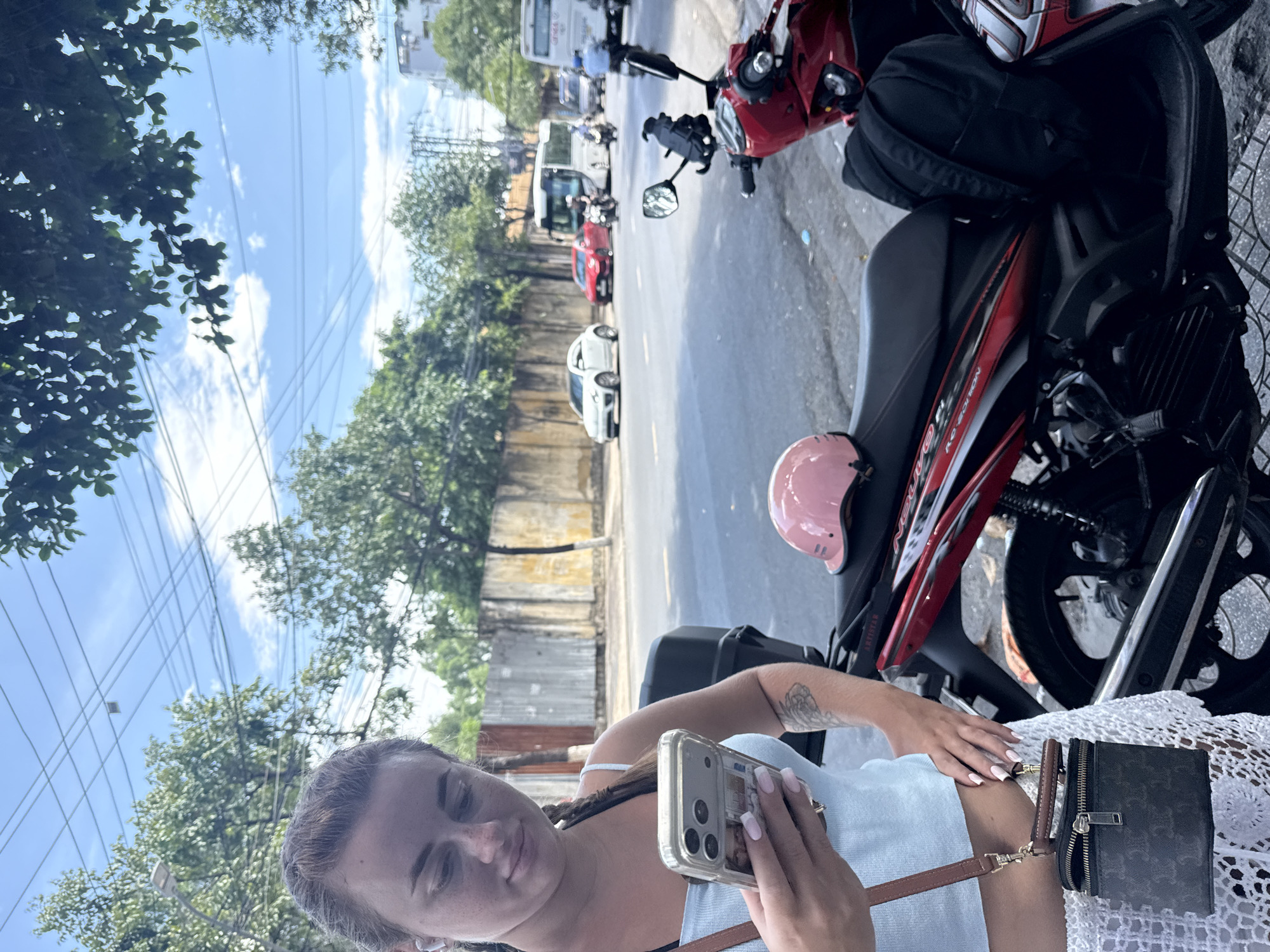 A young woman with braided hair and a light blue top is holding a smartphone and looking down at it. She is wearing white crochet shorts and a brown crossbody bag. A pink helmet rests on the seat of a red and black motorcycle next to her. The motorcycle has a black backpack behind the seat. In the background, a street in Nha Trang, Vietnam, is visible with traffic including cars, motorcycles, and buses. The environment is urban with trees and a concrete wall. The sky is blue with some clouds.