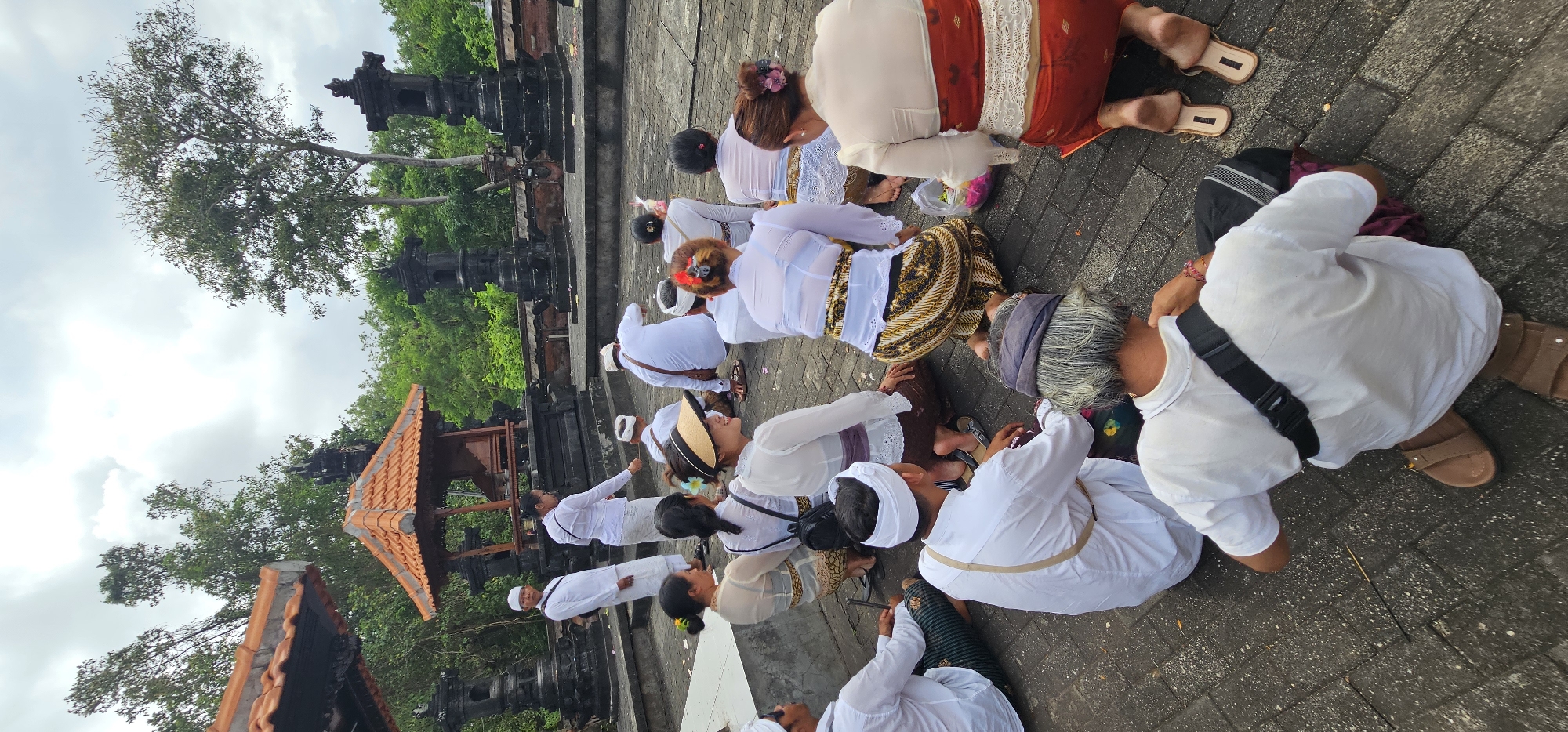 A medium, high-angle, outdoor shot, taken in the city of Kuta Selatan, Indonesia, shows a gathering of about a dozen people, most of whom are kneeling. The people are dressed in white shirts and skirts with patterns in golds, reds, and browns. In the background, there is a temple with black pillars and red-tile roofs, surrounded by green trees and a gray sky.