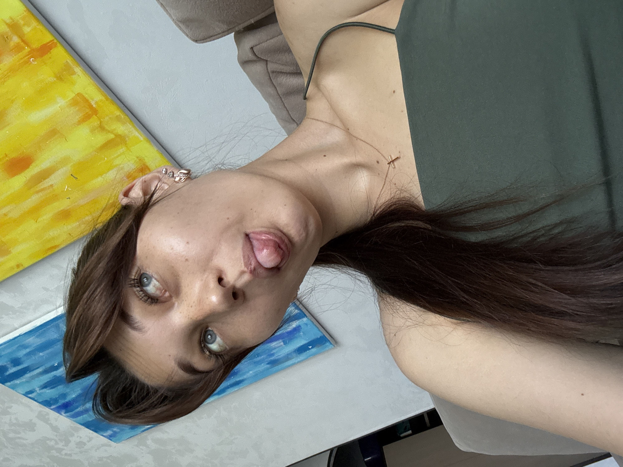 A young woman with brown hair pulled into a ponytail, and freckles on her nose and cheeks, is pictured from the shoulders up. She is wearing a green tank top and a rose gold necklace with a cross pendant. Her head is tilted upwards and to the left, with her eyes looking in the same direction. Her tongue is sticking out, and her mouth is slightly open. In the background, two abstract paintings are visible: one with blue and white brushstrokes, and another with yellow and orange brushstrokes. The woman appears to be indoors, with a couch visible to the right of the frame. There is no information in the image to determine the specific location in Novokuznetsk, Russia.