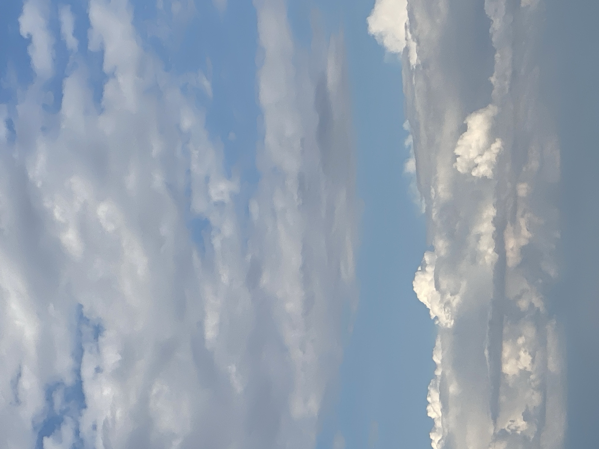 The image is a low-angle, full shot of a cloudy sky. The sky is a clear blue in the middle portion of the frame, with scattered white and gray clouds. Some clouds are wispy and thin, while others are thick and puffy, appearing to be cumulonimbus clouds. The bottom of the frame shows a hazy, grayish-blue area that could be the horizon or a distant landscape obscured by atmospheric conditions. There are no people, structures, or discernible landmarks visible in the image. The overall impression is of a natural, atmospheric scene with no specific urban or geographical features identifiable.