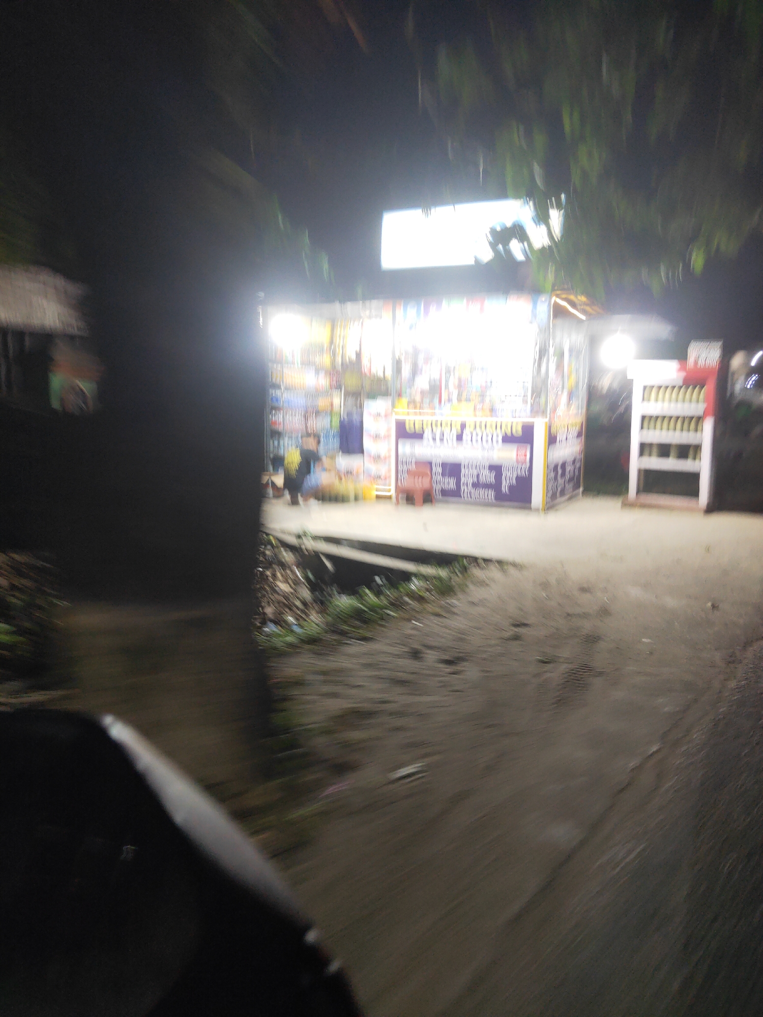 The image shows a roadside stall at night in Sintang, Indonesia. The stall is brightly lit, displaying various items for sale, including what appear to be snacks or small packaged goods. A person is visible inside the stall, and another figure is partially obscured in the foreground. The stall has a colorful facade with text in what seems to be a local language, possibly Indonesian, with some English words visible such as "BOOK". A red plastic chair is placed in front of the stall. Adjacent to the stall, a white shelving unit holds rows of bottles. The area around the stall appears to be a dirt path or road, with vegetation visible in the background and to the left. The overall impression is of a lively but somewhat informal marketplace at dusk or night.