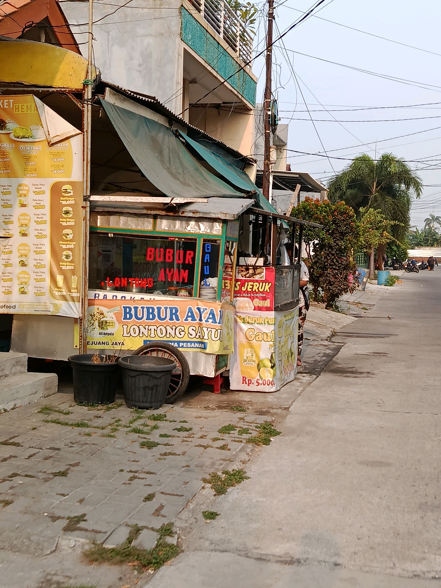 This image depicts a food stall in Bekasi, Indonesia, selling "Bubur Ayam" (chicken porridge) and "Lontong Sayur" (vegetable rice cakes). The stall is colorful, with a green awning and a prominent red sign displaying the food items. Various posters displaying food items and prices are also visible on the sides of the stall. Two large black planters and a bicycle wheel are placed in front of the stall. The stall is situated on a sidewalk next to a street with moderate traffic, including motorcycles. In the background, there are buildings, trees, and utility poles with many wires. The overall scene suggests a typical street-side food vendor in an urban environment.
