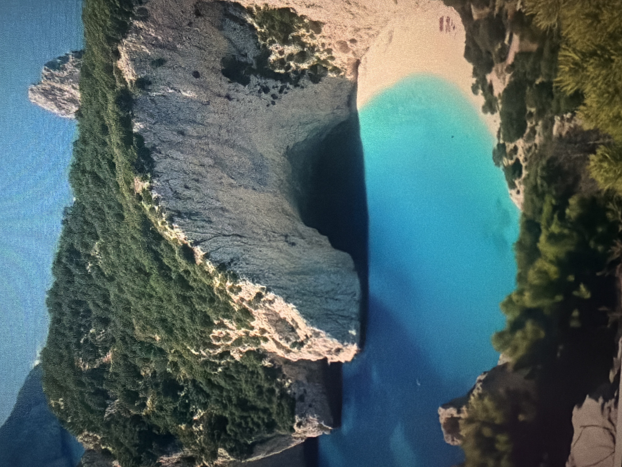 The image shows a scenic view of a cove with a sandy beach and turquoise water, framed by lush green hills and dramatic white cliffs. There is a large cave opening in the cliffs. The water appears calm and clear, with a gentle slope from the deep blue sea to the shallow turquoise shallows near the beach. The vegetation on the hills is dense and varied, suggesting a tropical or Mediterranean climate. The cliffs are rugged and weathered, with visible striations and darker patches indicating areas of shadow or different rock composition. The sunlight casts shadows that highlight the contours of the landscape. There are no people or man-made structures visible in the main part of the image. The overall impression is one of natural beauty and tranquility. The image does not show any recognizable elements of Baan Mae Nam, Thailand.