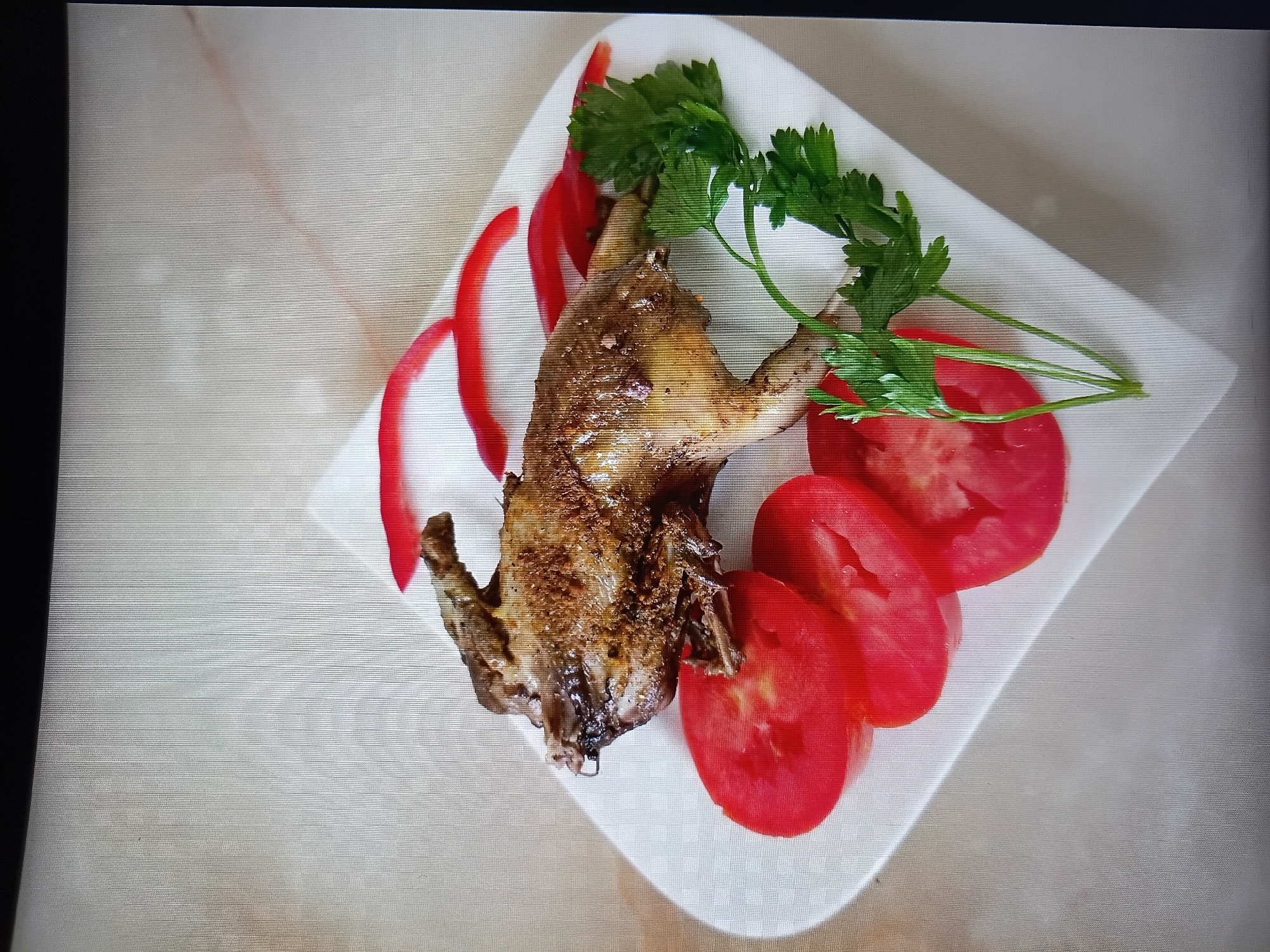 A roasted quail, garnished with sliced tomatoes, red bell pepper strips, and parsley sprigs, is presented on a white, square plate. The plate rests on a textured, light-colored surface, possibly a tablecloth or countertop. The lighting suggests an indoor setting, with shadows cast by the food and plate onto the surface. The overall composition is a top-down view of the dish. There is no information in the image to indicate a location in Tiraspol, Moldova.