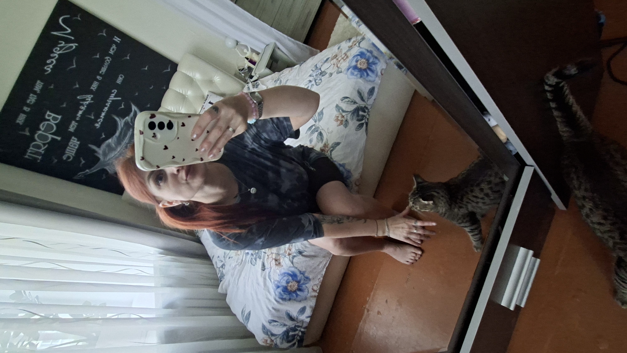 The image is a mirror selfie taken in a bedroom in Myadzyel, Belarus. A woman with red hair pulled back in a ponytail is sitting on the edge of a bed. She is wearing a dark tie-dye t-shirt and black shorts. She is holding a smartphone in her right hand, taking the picture. A cat with tabby markings is sitting on the floor in front of the bed, looking up at the woman. The bed is covered with a floral-patterned comforter. In the background, there is a black headboard with white writing and a drawing of a feather. The room has wooden floors and sheer curtains covering a window. There is a large dresser with a mirror in the foreground, reflecting the scene.