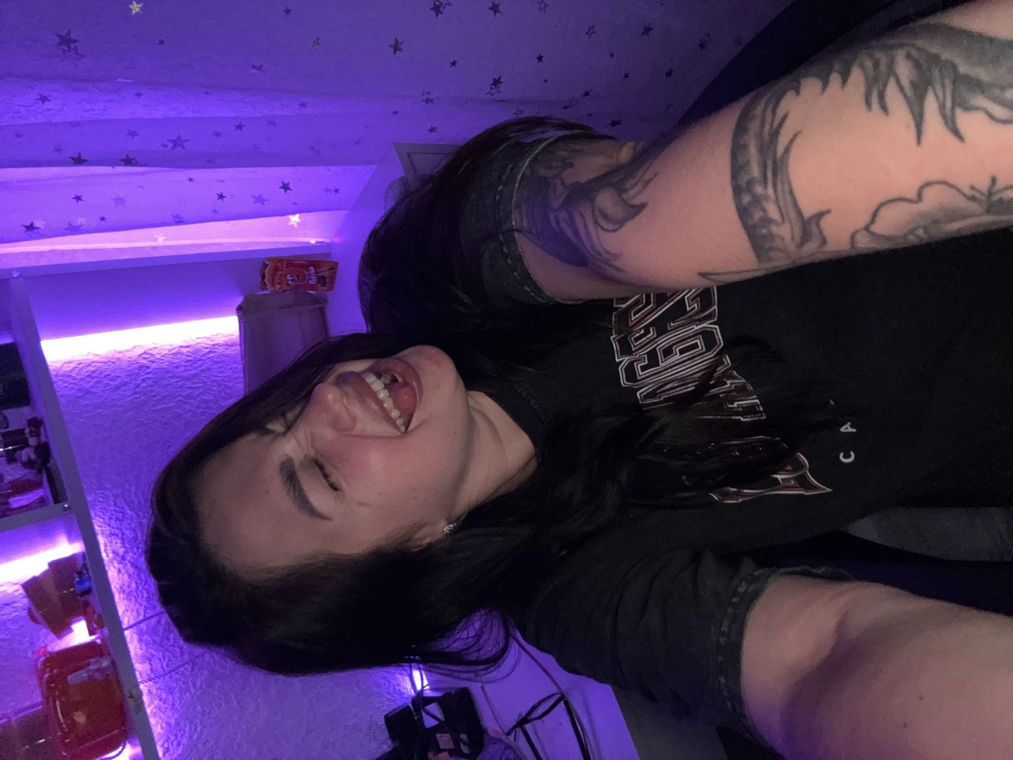 The image shows a young woman with dark hair, laughing with her eyes closed and mouth open. She is wearing a black t-shirt with white lettering and has a tattoo on her right arm. The background is dimly lit with purple and pink lighting, with shelves holding various items and a curtain with star patterns. There is no information in the image or its crops to indicate the location in the city of Tolyatti, Russia.