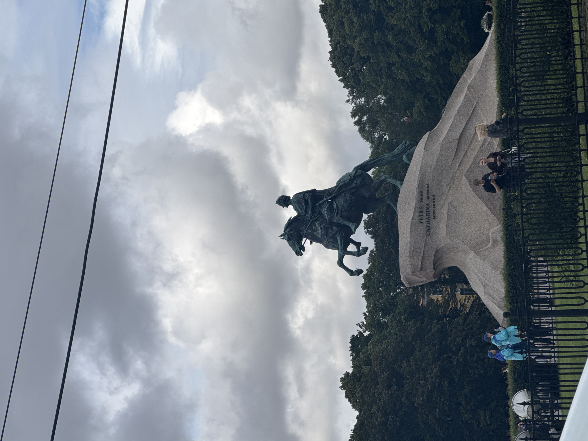 The image features a dramatic equestrian statue of a man on horseback, likely a historical figure, positioned against a cloudy sky. The statue is dark bronze and appears to be in motion. To the right of the statue is a large, rough-hewn granite base with text inscribed on its side. The text reads "PETRO PRIMO CATHARINA SECUNDA" and "MDCCLXXXII". This suggests the statue is of Peter the Great and possibly related to Catherine the Great, with an inscription dating to 1782.
Below the granite base, a black metal fence encloses a grassy area with trees in the background. Several people are visible in the middle ground, near the fence. Some are standing, while others are seated. Two individuals in the foreground are wearing bright blue jackets, suggesting they might be tourists or part of a group. The lighting and clouds indicate it could be daytime, possibly overcast or nearing dusk. The overall atmosphere is solemn and grand due to the imposing statue and its historical inscription.