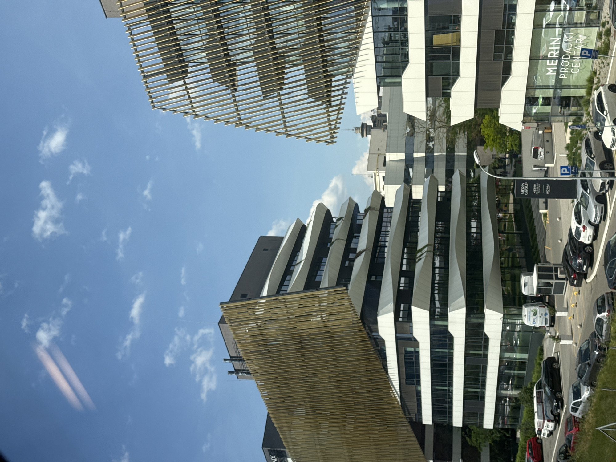 The image depicts a contemporary architectural scene in Belgrade, Serbia. The angle of the photograph is unconventional, showcasing buildings tilted into the blue sky. The architectural elements include modern buildings with glass facades and decorative horizontal and vertical striped lines. In the background, a commercial sign reading "MERIN Prodajni Centar" is visible, along with parked cars and some greenery. The sky is clear with scattered white clouds.