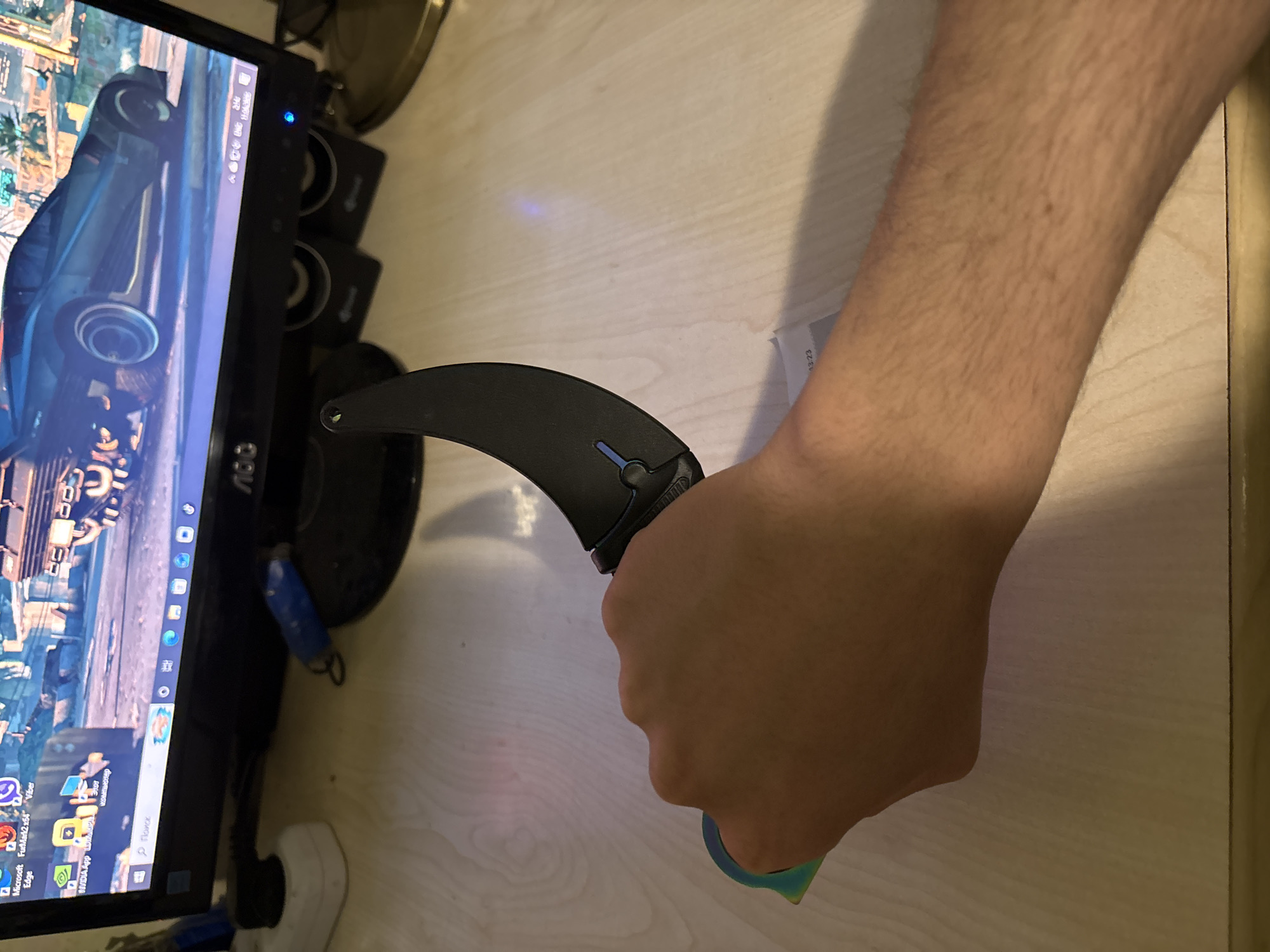 A close-up, slightly angled shot shows a person holding a karambit knife in their right hand, with the blade facing towards the left. The hand and forearm are pale, with noticeable hair. In the background, a computer monitor displays a game, and there are speakers behind the knife. The surface beneath appears to be a light-colored desk or table. There is nothing in the scene that would indicate Yerevan, Armenia.