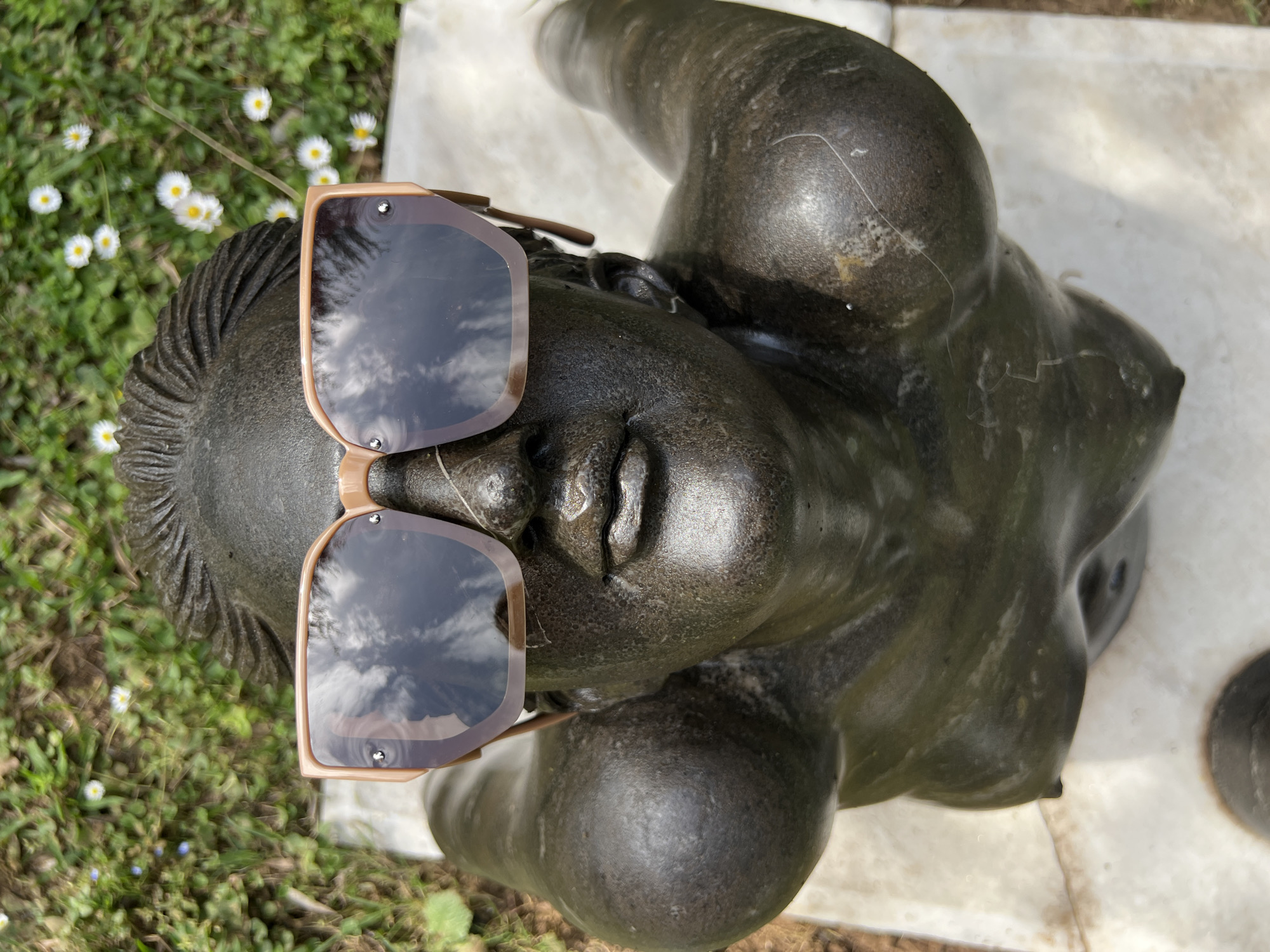 An eye-level shot features a dark-colored, seemingly bronze bust of a woman wearing sunglasses. The sunglasses have a tan frame and reflect a sky with clouds in their lenses. The bust has detailed hair and facial features, with an obscured view of the body. The bust rests on a white marble surface with hints of texture. To the left of the bust, there is a patch of green grass with small white flowers. The setting may be a park or garden, likely located in Petrovac na Moru, Montenegro. The composition focuses on the bust and its immediate surroundings.