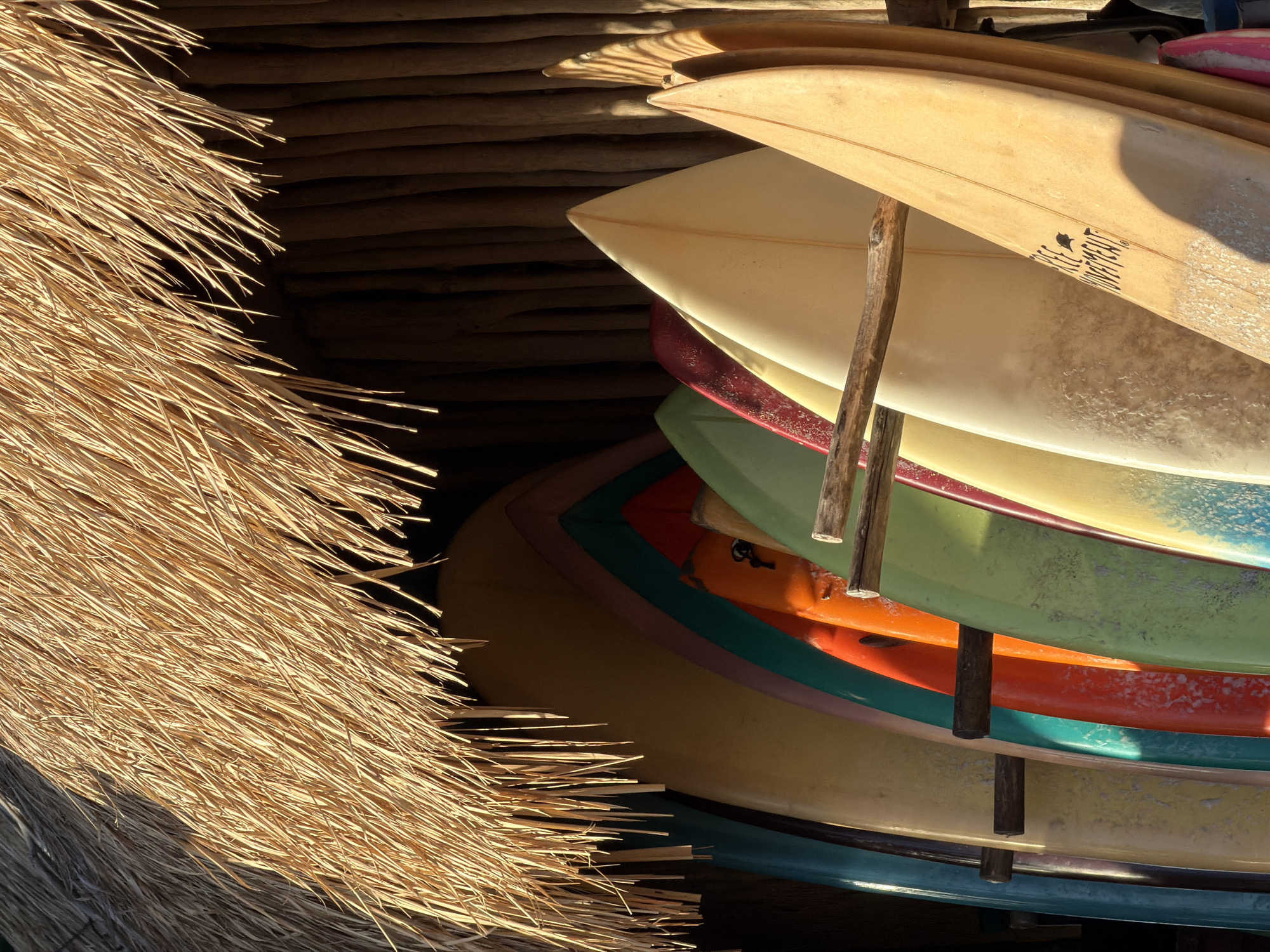 A stack of surfboards leans against a wall under a thatched roof in North Kuta, Indonesia. The surfboards are in varying colors, including orange, green, yellow, and red. The topmost surfboard is cream-colored and bears a logo. Two vertical, rough-hewn poles support the surfboards. The wall behind is dark, possibly made of stacked timber. The roof is made of straw-like material, filling the left side of the frame.