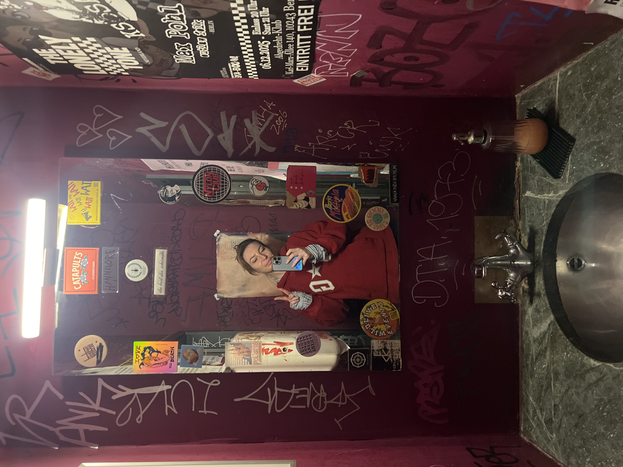 A young woman is reflected in a graffiti-covered mirror in a bathroom. She is holding a smartphone and posing for a photo. The bathroom has burgundy walls covered in graffiti, stickers, and posters. A flyer indicates this location may be in the Karl-Marx-Allee district, or possibly a club in Berlin, Germany. A stainless steel sink and a stone countertop are visible.