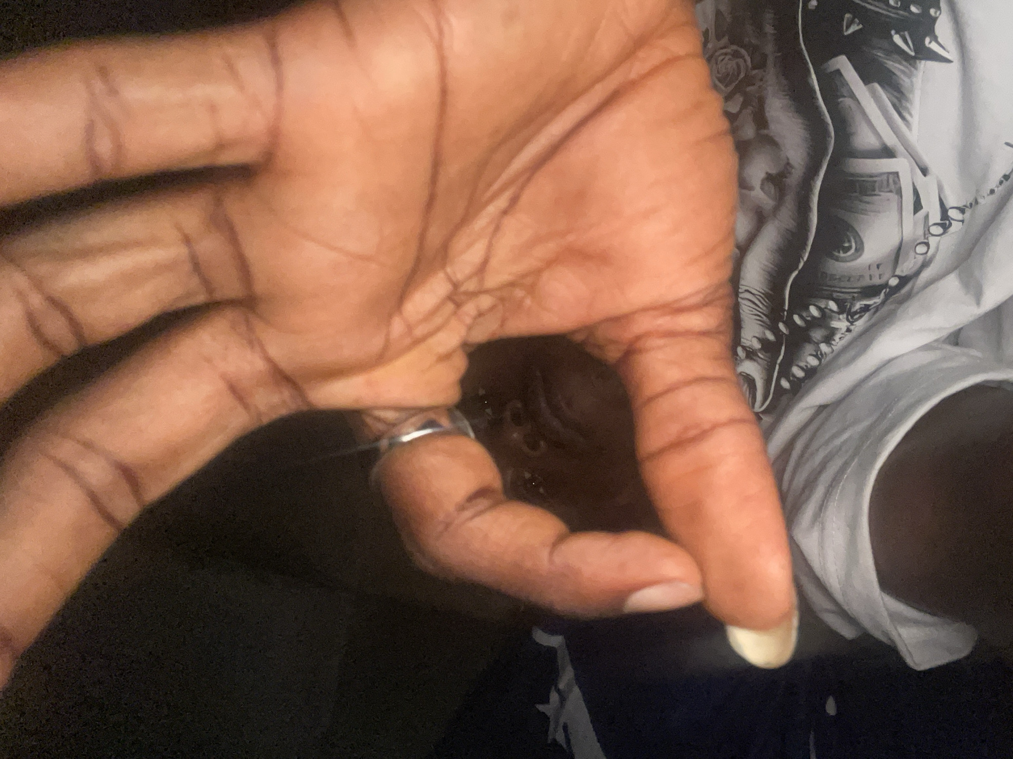 An overhead shot captures a dark-skinned hand with visible wrinkles and a silver ring on the ring finger. The hand is extended towards the right side of the frame. The background includes a white t-shirt with a printed graphic, partially visible, and dark clothing. The location is unknown, and there is no visible reference to Lokoja, Nigeria.