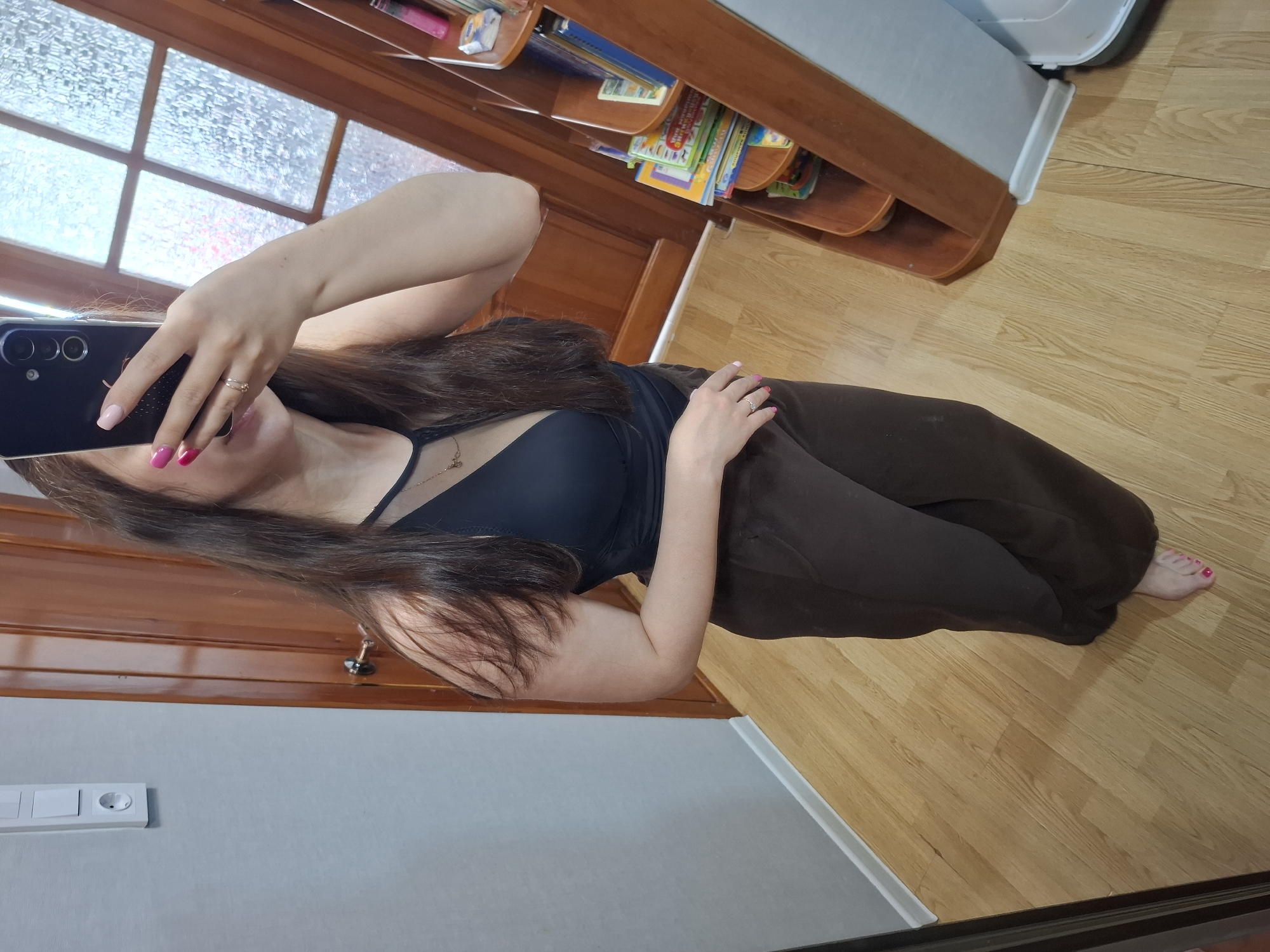 A person is taking a selfie indoors, likely in a residence. The individual is wearing a black tank top and dark brown sweatpants, and has painted their fingernails and toenails pink. The phone being used for the selfie is black with multiple lenses on the rear. In the background, a window with a divided grid pattern is visible, along with a wooden bookshelf filled with books and other items. The floor is made of light-colored wood. A light switch can be seen on the wall. The location is unspecified and is not apparent from the information provided. The city of Chaikovsky, Russia cannot be determined from the provided image.