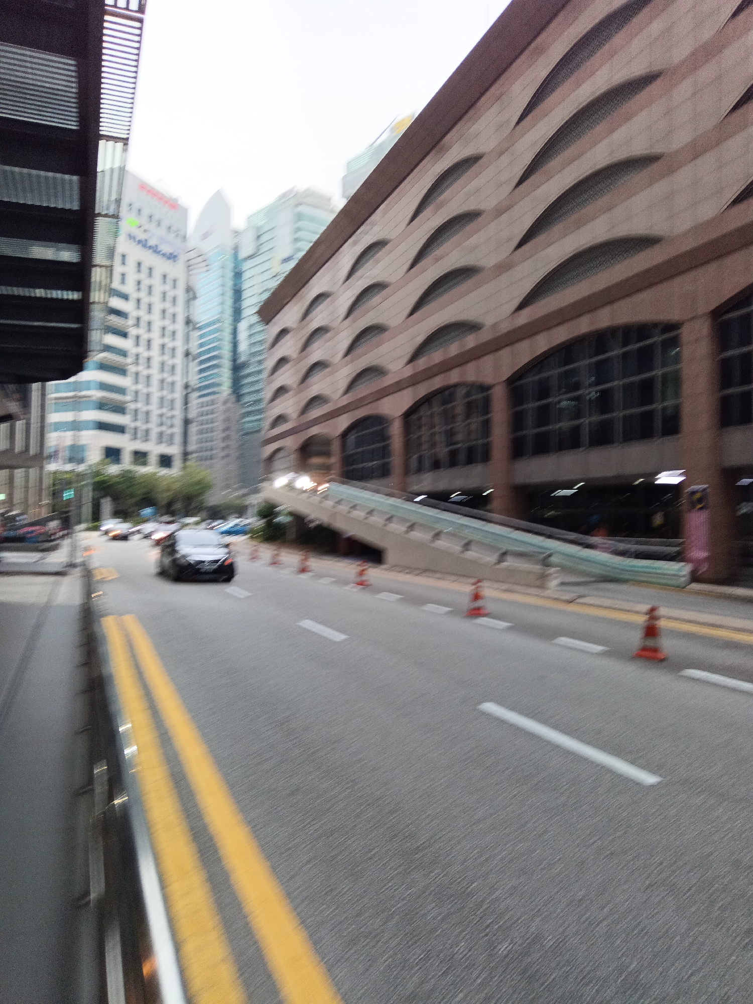 This is a slightly blurred, eye-level, wide shot of a city street in Kuala Lumpur, Malaysia, on an overcast day. The perspective is from a moving vehicle, looking forward and slightly to the right. In the foreground, the asphalt road features double yellow lines on the left and white lane markings. A metallic barrier is visible on the far left.
Dominating the right side of the frame is a large, multi-story building with a reddish-brown facade and distinctive arched window designs. To the left of this building, there is an elevated pedestrian walkway or ramp leading into the building. Orange traffic cones are placed along the road.
In the middle ground, several cars are visible on the road, including a black sedan in motion and other vehicles further down the street. The background is filled with various tall buildings, including office towers and other urban structures, some appearing to be hotels or commercial buildings. The sky is uniformly grey, indicating cloudy weather. The overall impression is of a busy urban environment during daylight hours.