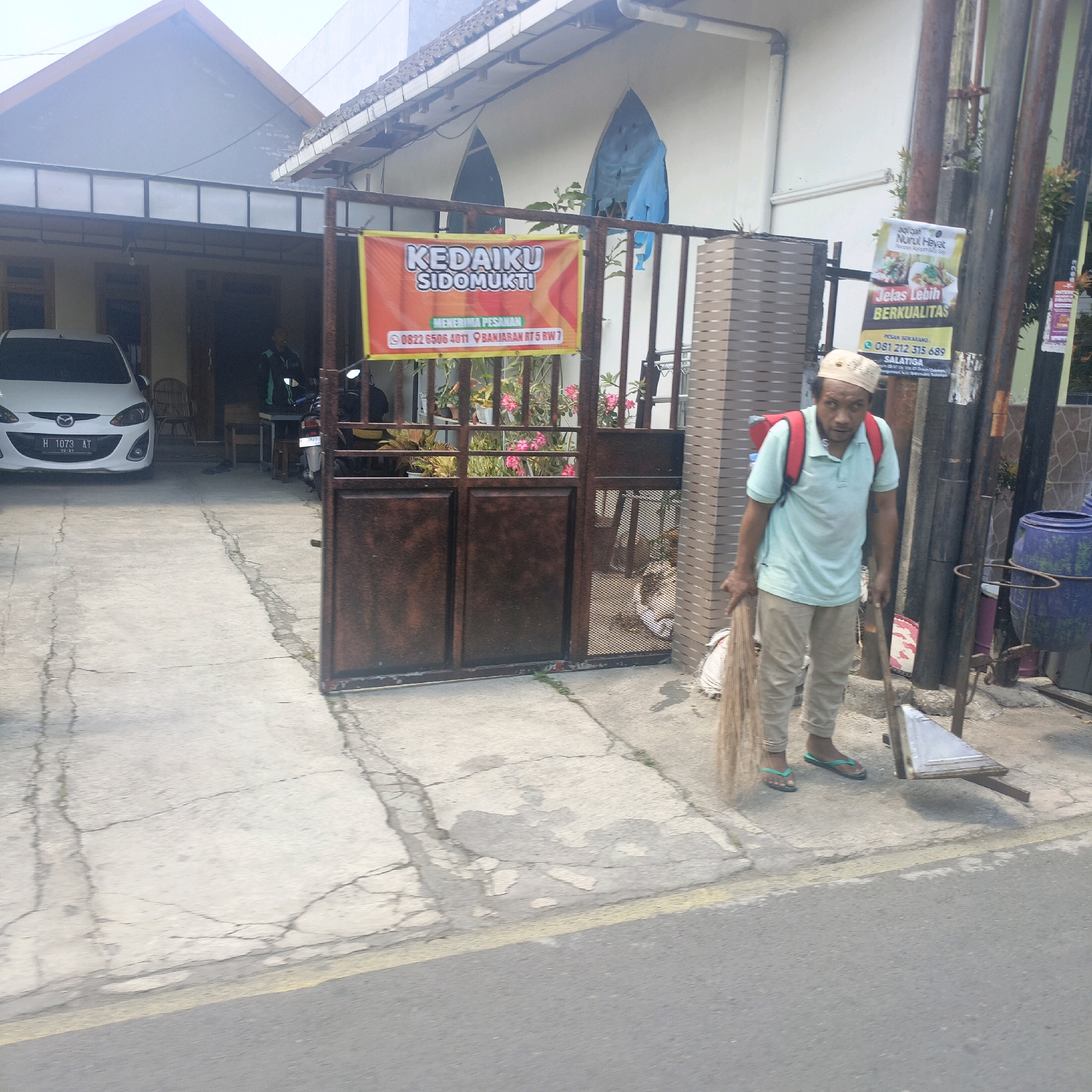 A man, wearing a light blue polo shirt, beige pants, a patterned cap, and green flip-flops, is sweeping the sidewalk with a broom. He has a red backpack on. Behind him is a building with a brown gate. A white car with the license plate H 1073 AT is parked in front of the building. A red and yellow banner with Indonesian text is hanging on the gate. To the right of the man is a large blue trash can. The scene appears to be on a street in Salatiga, Indonesia.