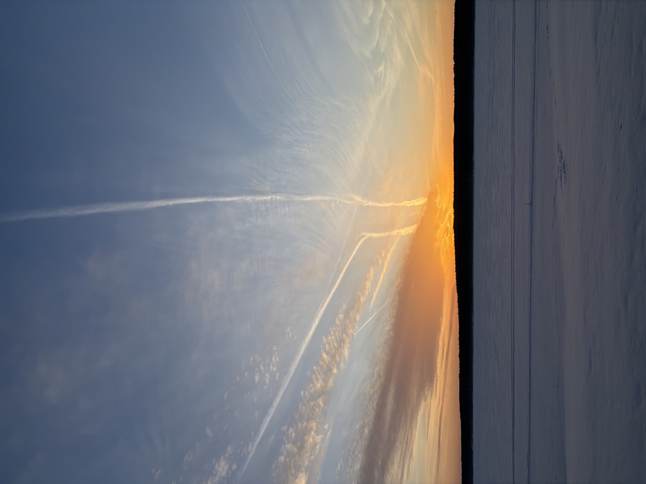 The image displays an outdoor winter landscape during a sunset or sunrise, situated in Chaikovsky, Russia. The foreground consists of a vast, flat expanse of snow-covered surface, likely a frozen body of water, occupying the lower third of the frame. Faint, parallel linear depressions are discernible across the snow, suggesting tracks. A dark, dense treeline forms a horizontal silhouette along the horizon, separating the snow from the sky. The sky, dominating the upper two-thirds of the image, exhibits a gradient of colors, with vibrant orange and yellow hues concentrated near the horizon, transitioning upwards through shades of pink and light blue to a deeper blue at the top. Multiple aircraft contrails are visible. Two distinct, parallel contrails ascend vertically from the horizon near the center-right. Several other contrails, some broader and more dissipated into wispy cirrus clouds, traverse the sky diagonally and horizontally from the left, converging towards the bright horizon. No human or animal subjects, explicit actions, or artificial structures other than the atmospheric contrails are present in the scene.