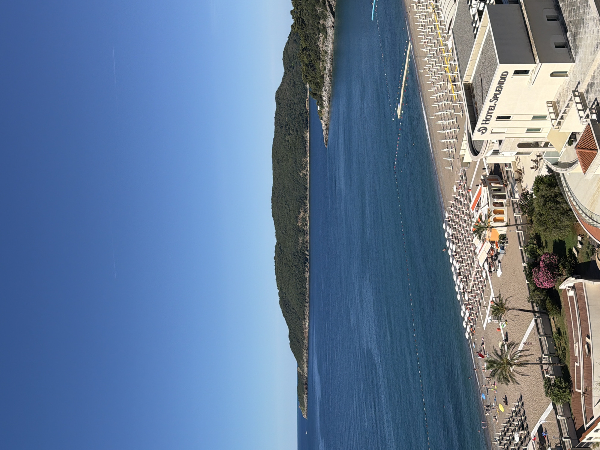 The image shows a clear, bright, sunny day with a vast expanse of blue sky above a deep blue sea. In the distance, a lush green, tree-covered island or peninsula juts out into the water, with a rocky shoreline. The mid-ground features a sandy beach lined with rows of white umbrellas and lounge chairs. Several people are visible on the beach, some walking, some sitting under umbrellas. Palm trees dot the sandy area, and there are some small buildings with orange roofs. In the foreground, a modern, multi-story building with the prominent sign "HOTEL SPLENDID" is visible. This hotel appears to be situated on the coast, overlooking the beach and the sea. The overall scene depicts a popular resort destination, likely a beach hotel in Boreti, Montenegro, with calm waters and a clear sky.
