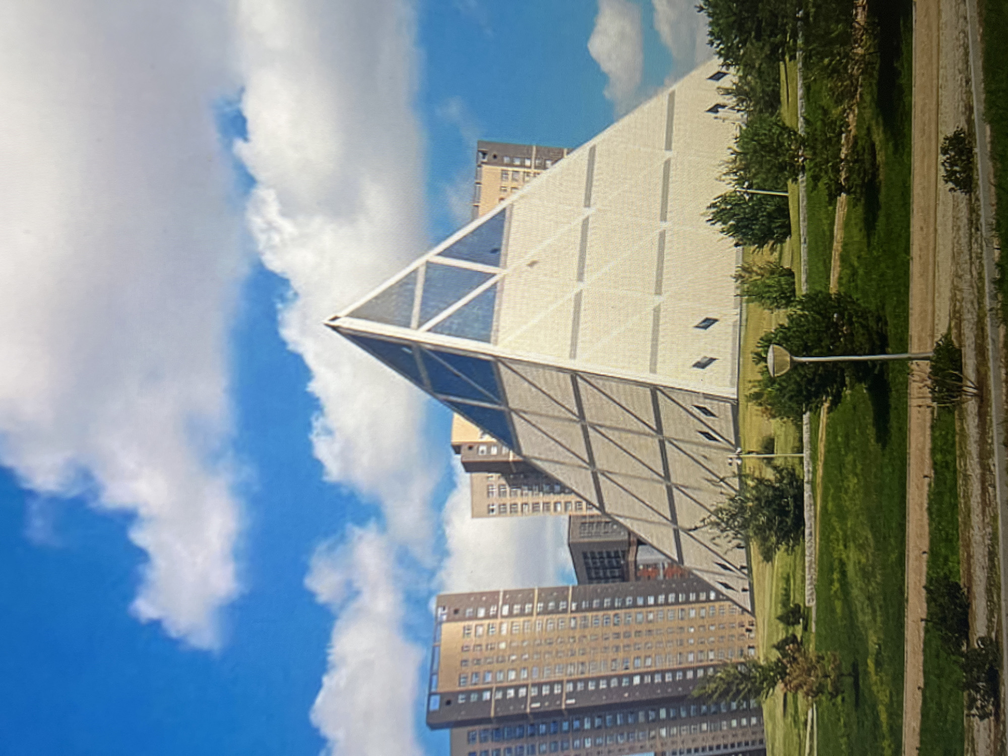 The image shows a large pyramid-shaped building with glass and light-colored facades, situated in a grassy park area. Several tall, modern apartment buildings with many windows are visible in the background, to the left of the pyramid. The sky is blue with scattered white clouds. The ground in front of the pyramid is a gently sloping lawn with scattered trees and bushes. A paved path runs through the foreground, with a lamppost on the right side. The scene appears to be a public space in a city, likely in Kazakhstan, given the mention of Qarağandı city administration. There are no people visible in the image.