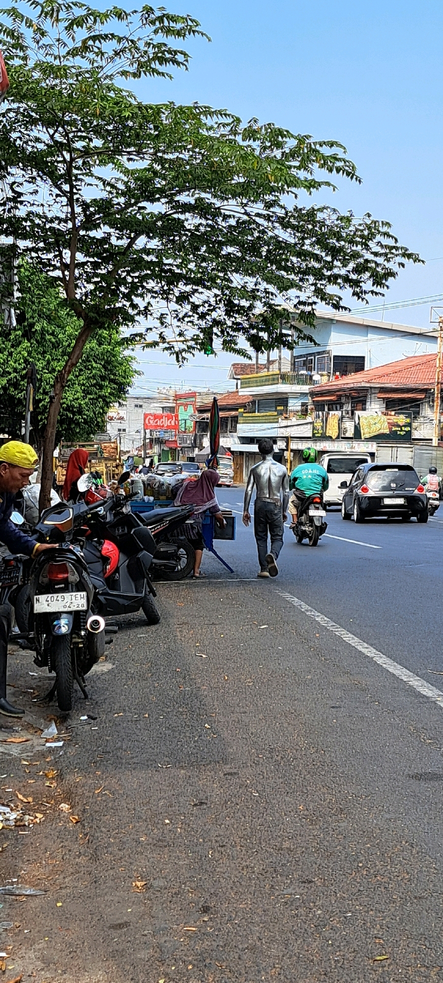 An outdoor scene on a street in Petungasri, Indonesia, depicts a street with several motorbikes parked along the side and a cart laden with goods. A man in a yellow cap is seen near the motorbikes, partially obscured by a tree. In the background, several buildings line the street, some with red-tiled roofs. Two individuals are walking away from the camera on the street. One is a person painted silver from head to toe, wearing dark pants and sneakers. The other is wearing a green shirt with "GO JEK" written on it and riding a motorbike. A black car is driving away from the scene, and another motorbike with a rider is further down the road. The sky is clear and blue.