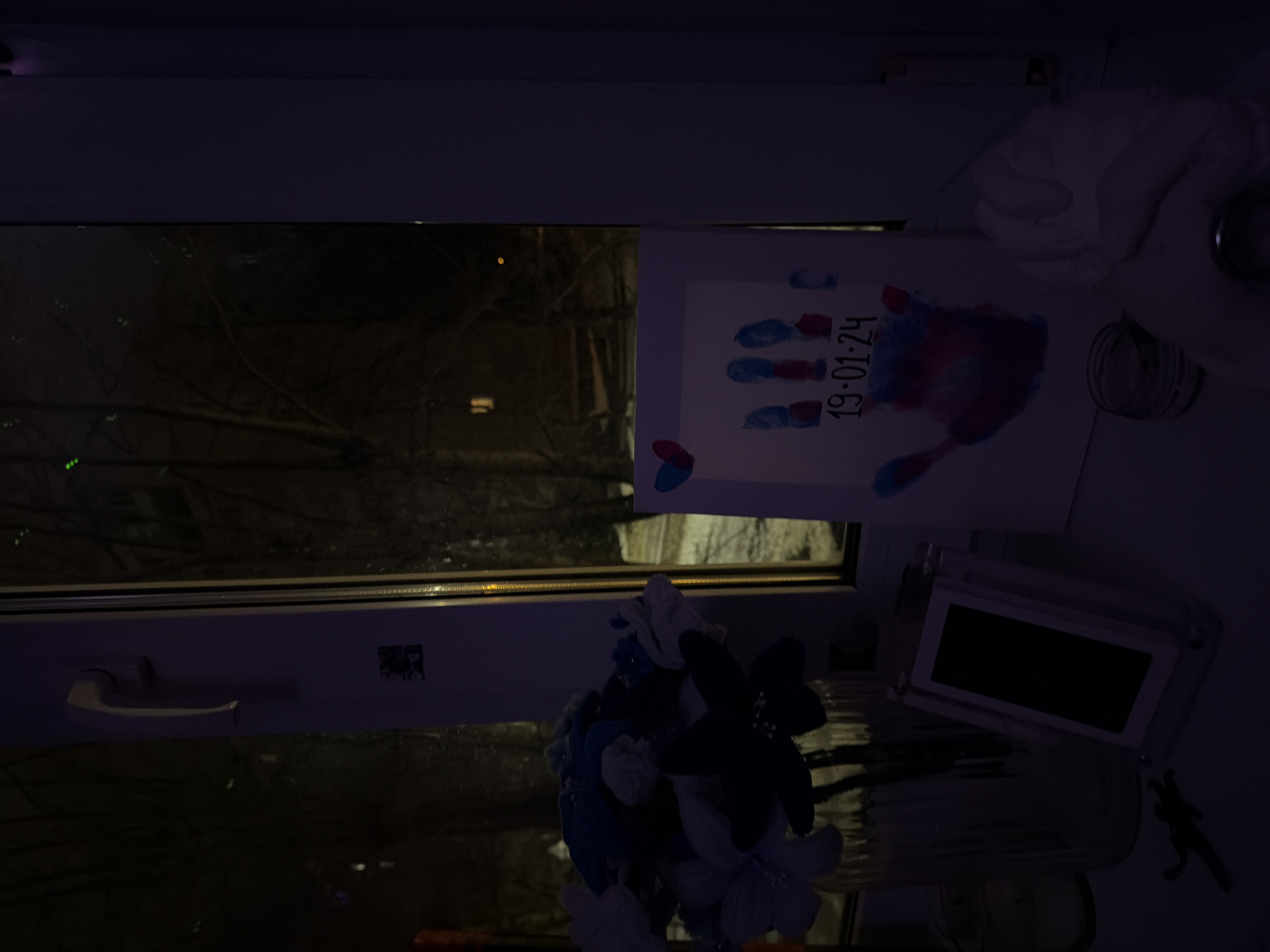 The image depicts a window in what appears to be a dark room. Outside the window, bare tree branches are visible, indicating a possible winter season. Inside, a handmade print featuring a child's handprint and the date "19.01.24" is propped up against the window. Also present on the window sill are a vase with artificial blue and black flowers, a small digital photo frame with a dark screen, and what appears to be a container, possibly glass. A portion of a white curtain is visible on the right side. The handle of the window is on the lower left side. There are no people visible in the window reflection. The scene is dimly lit, making it difficult to discern finer details. Based on the date format used on the handprint (day.month.year), it is plausible that this image was taken somewhere in Europe or Russia. Given the additional context that this is in Perm, Russia, this observation is very likely correct.