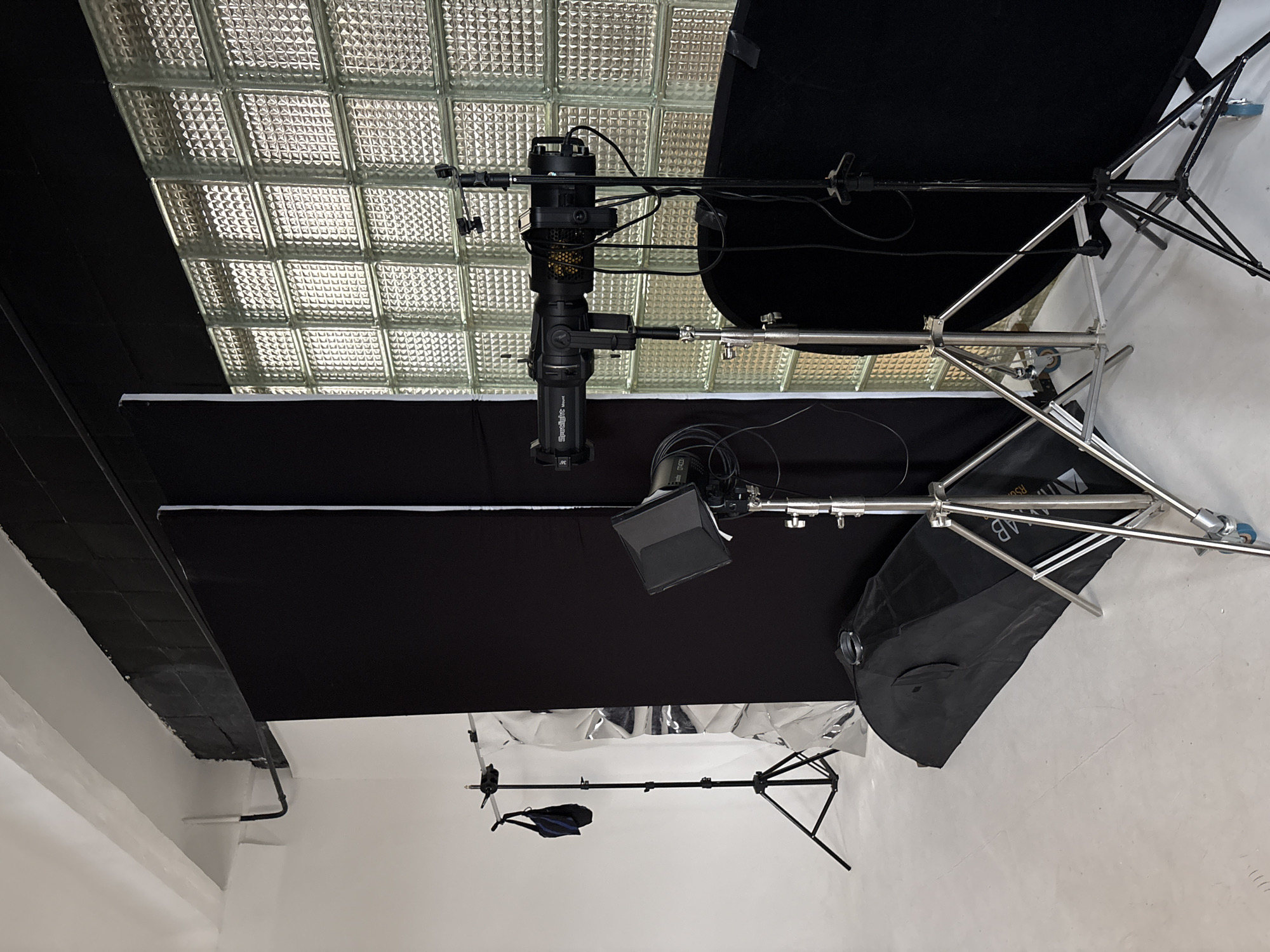 The image depicts a photography studio setup. The room has white walls and a ceiling with glass brick panels. A black backdrop is suspended, supported by stands. Various lighting equipment, including studio lights and softboxes on stands, are positioned around the backdrop. Cables are visible. The image does not include people. There is no indication that this is taken in Veliky Novgorod, Russia.