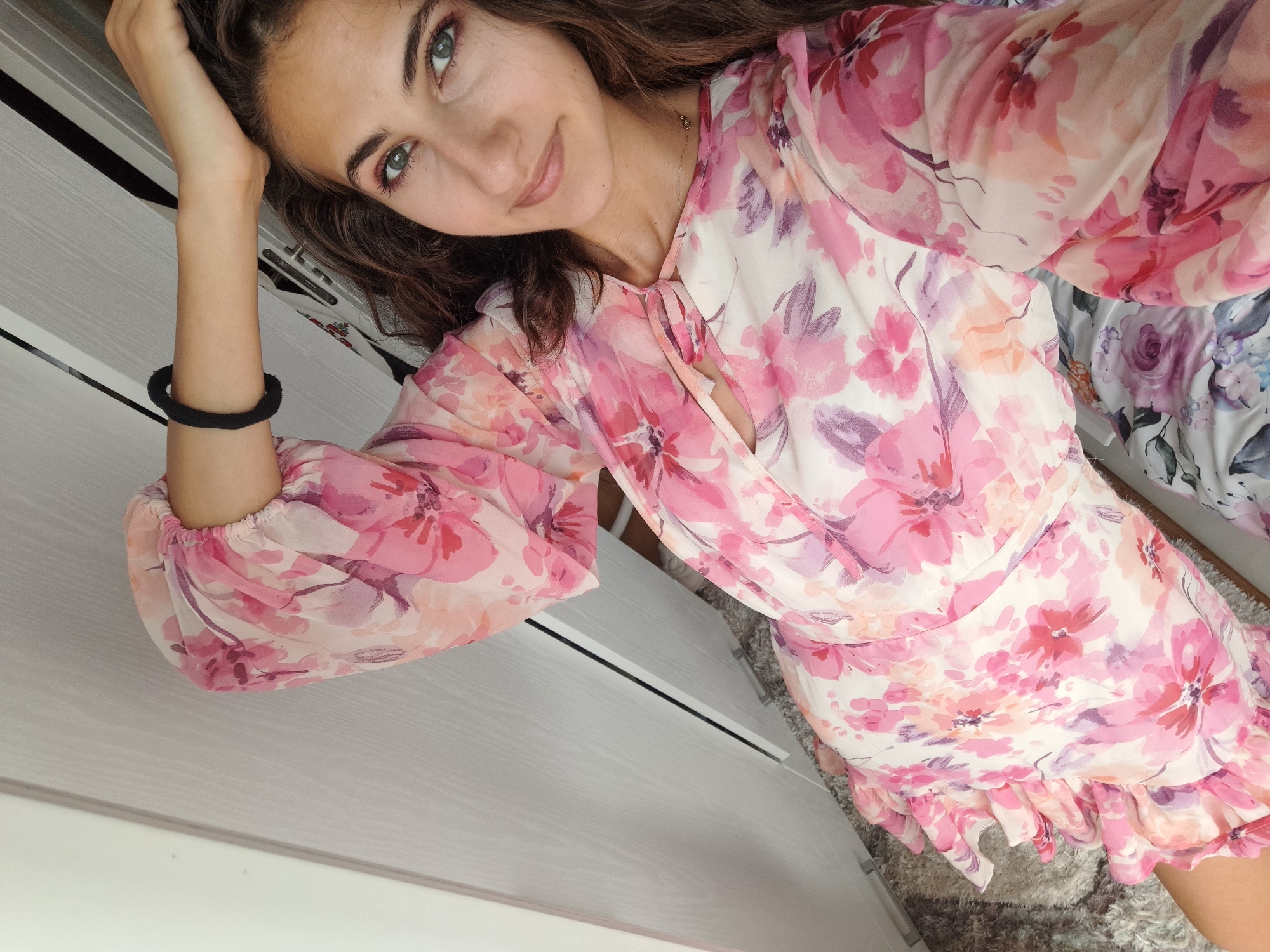 A young woman with long, wavy brown hair and striking green eyes is captured in a selfie. She is wearing a flowing pink and white floral print dress with a V-neck and tie detail. Her left arm is bent, with her hand resting on her head. She has a thin black bracelet on her left wrist. Her expression is relaxed and she is smiling softly at the camera, conveying a sense of gentle happiness. The background shows a hint of a grey, textured surface, possibly a dresser or cabinet, with a carpeted floor visible in the lower right corner. The lighting appears to be natural, suggesting it could be daytime. The overall impression is one of casual elegance and youthful charm.