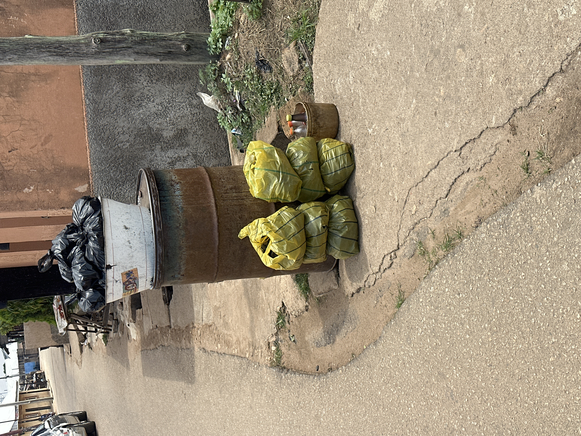 In an outdoor urban setting, likely on a street or sidewalk, several items are piled on the ground. A large, rusted metal barrel stands upright, topped with a white bucket overflowing with black trash bags. To the right of the barrel, a stack of three bright yellow plastic bags is propped against it. Next to the yellow bags is a small, brown bucket containing several bottles with colored caps. The ground appears to be a mixture of asphalt and concrete, with some areas of dirt and sparse vegetation. In the background, a wall made of rough, textured material is visible, with a weathered wooden pole standing to the right of it. A vehicle is partially visible on the left side of the image, suggesting a street environment. The overall scene depicts refuse and discarded items in a seemingly public area.