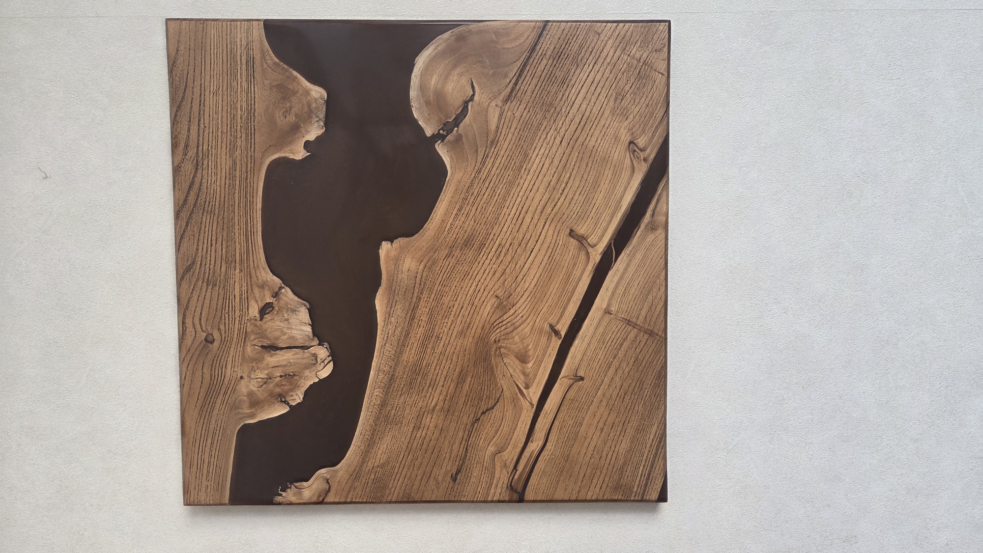 A square wooden slab featuring a large, irregular river of dark brown resin runs through its center. The wood grain is visible, and knots are incorporated into the piece. A dark, thin line of resin cuts diagonally through the wood on the right side. The slab is mounted on a light-colored wall. Based on location information, the image was taken in or around the city of Chaikovsky, Russia.