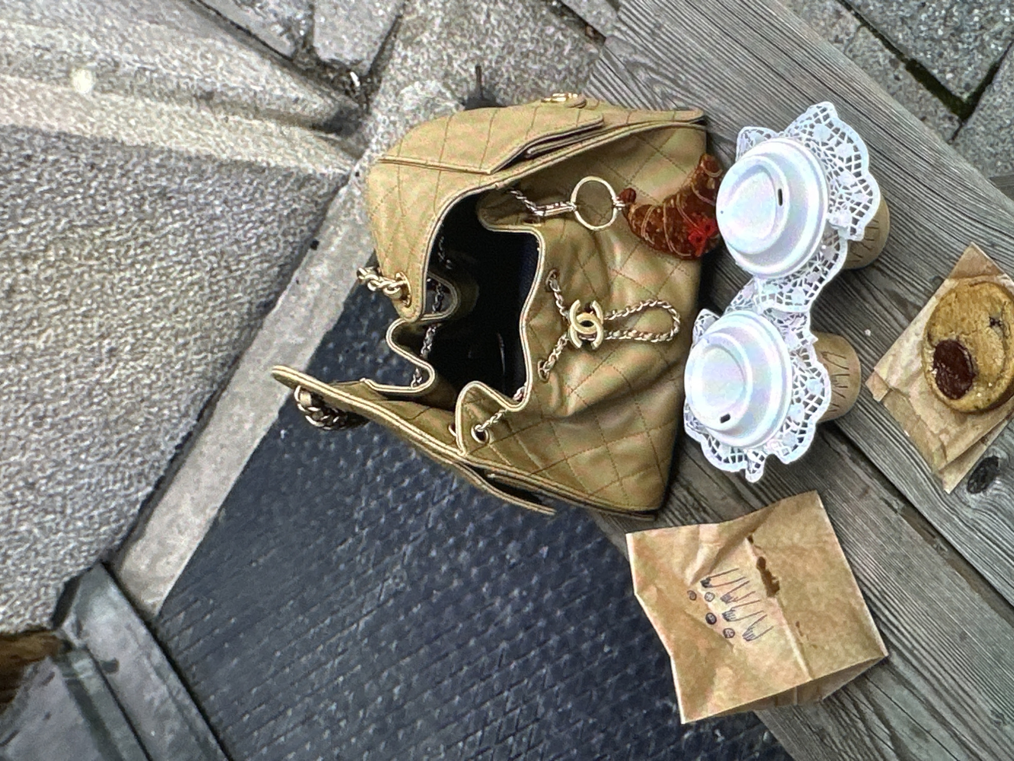 The scene shows a beige Chanel bucket bag, two cups of coffee with white lace doilies around them, a cookie in a paper bag, and a brown paper bag with drawings on it, all sitting on a weathered wooden surface. The surface is likely part of an outdoor table or bench. Adjacent to the wooden surface is a dark, textured surface, possibly a paved area or sidewalk, and a lighter gray stone surface. The scene evokes a casual outdoor setting, perhaps a cafe or park in Yerevan, Armenia, where someone has placed their belongings while taking a break.
