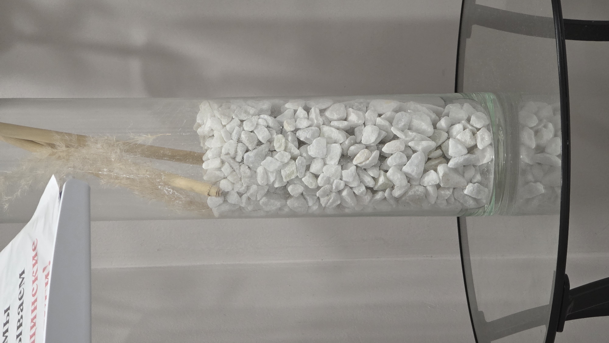 The image displays a clear glass cylindrical container, oriented horizontally, filled with decorative elements. The right two-thirds of the container are densely packed with numerous small, irregularly shaped white pebbles or stones. To the left of the pebbles, several dried, light brown reeds or pampas grass stalks are arranged within the container, with some feathery parts extending into the pebble section.
The container appears to be resting on or near a modern glass-topped table, a portion of which, including its black frame, is visible on the right side of the image. The background is a plain, light-colored wall or surface.
On the far left, a white object, possibly a sign, book, or brochure, is partially visible, obscuring a small part of the container. This object features red Cyrillic text. Visible fragments include "МЫ" (My - We), "ЫВАЕМ" (yvaem - part of a verb like "invite" or "deliver"), and "ИЦИНСКИЕ" (itsinskiye - part of a word like "medical" or "medicines"). The overall scene suggests an indoor decorative arrangement. No people or activities are present.