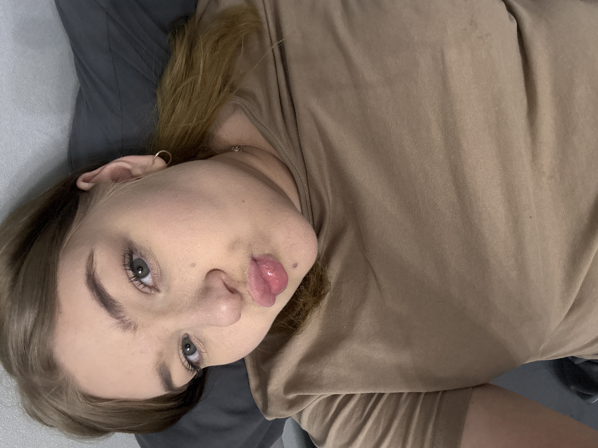A fair-skinned woman with light brown hair is lying down. She has gray eyes and is wearing an earring in her left ear. She is wearing a brown t-shirt and is lying on a gray surface. The location is unknown.