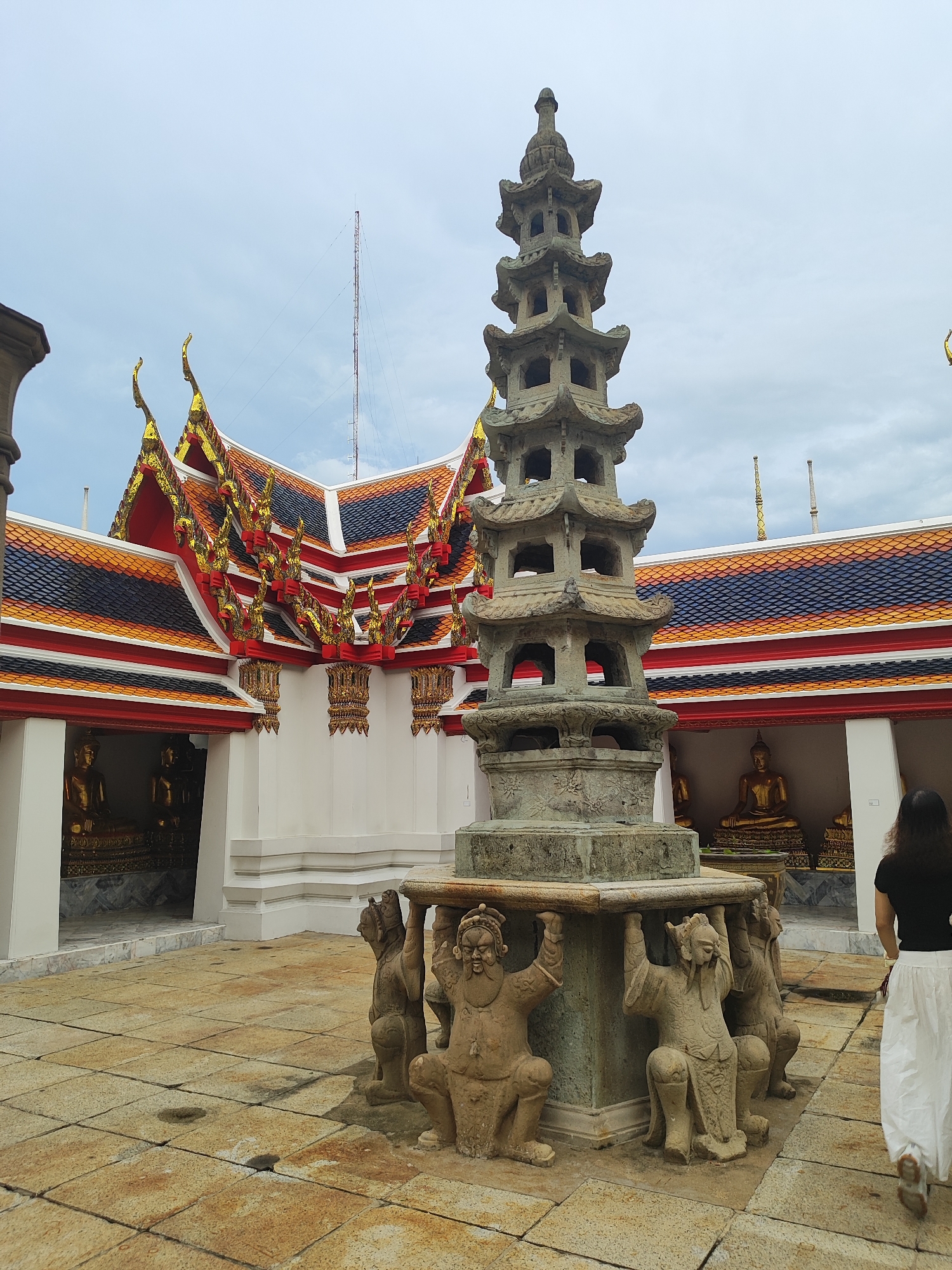 A stone stupa is positioned in the foreground of the image, supported by four carved figures. The stupa has multiple tiers, with arched openings on each level. The figures supporting the stupa are in a crouched position, holding the structure above them. The ground is paved with square tiles, some of which appear wet.
Behind the stupa, a white building with a red and gold tiled roof is visible. The roof has ornate decorative elements. Several golden Buddha statues are housed in arched alcoves along the building's facade.
A woman is walking away from the camera on the right side of the image. She has long, dark hair and is wearing a black t-shirt and a long white skirt.
The sky is overcast with a few visible antennas on the horizon. The scene appears to be at a temple in Bangkok, Thailand, likely Wat Pho, known for its reclining Buddha and numerous stupas.