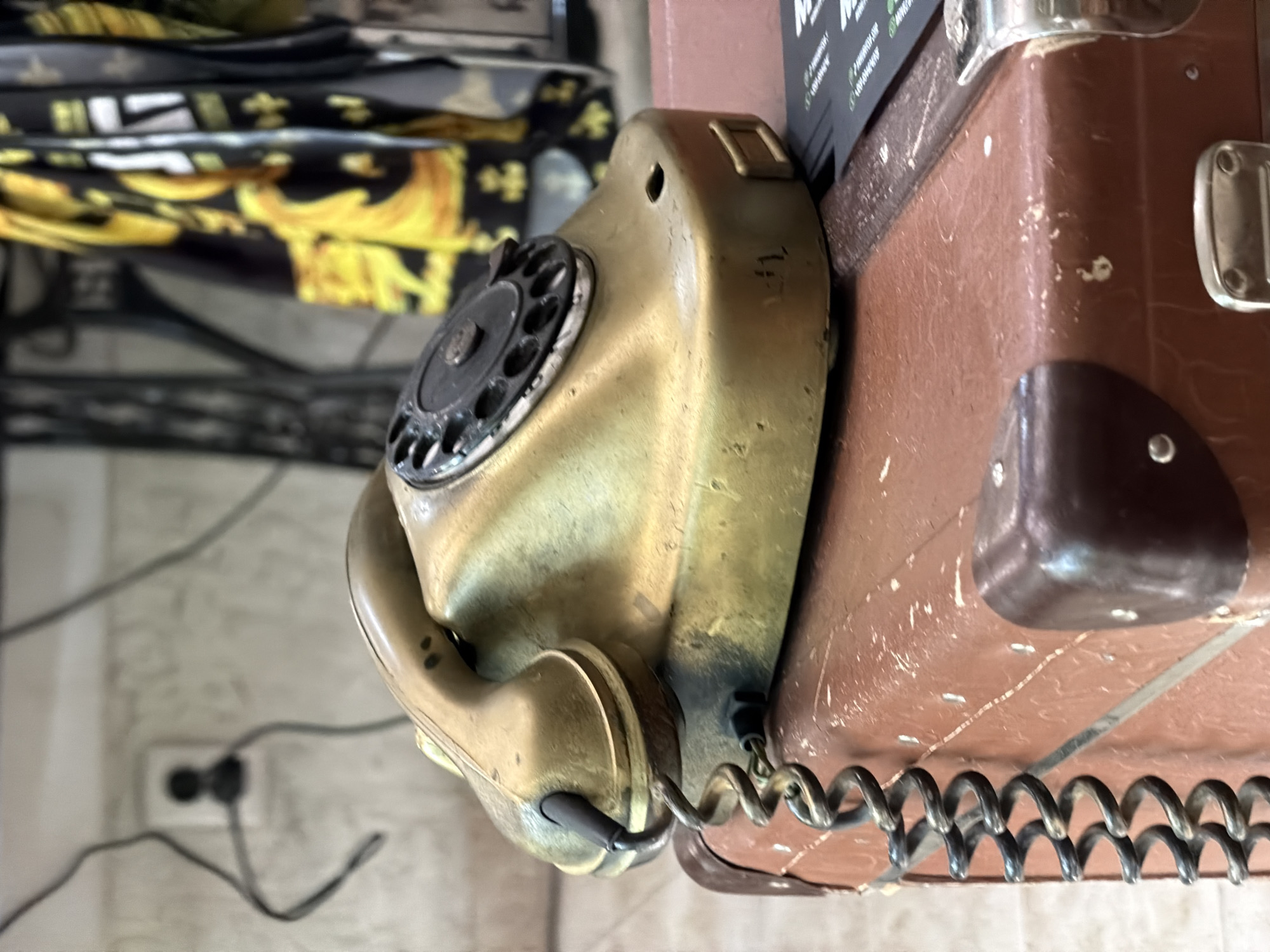 A close-up captures a vintage rotary dial telephone mounted on a brown, possibly leather-clad, object. The phone is a tarnished brass or gold color, showing signs of age and use. The coiled handset cord is connected to the phone's base. In the background, out of focus, there's a patterned item, possibly clothing, and a glimpse of a power outlet and wiring on a wall. Specific location within Sofia, Bulgaria is not discernable from the given image.