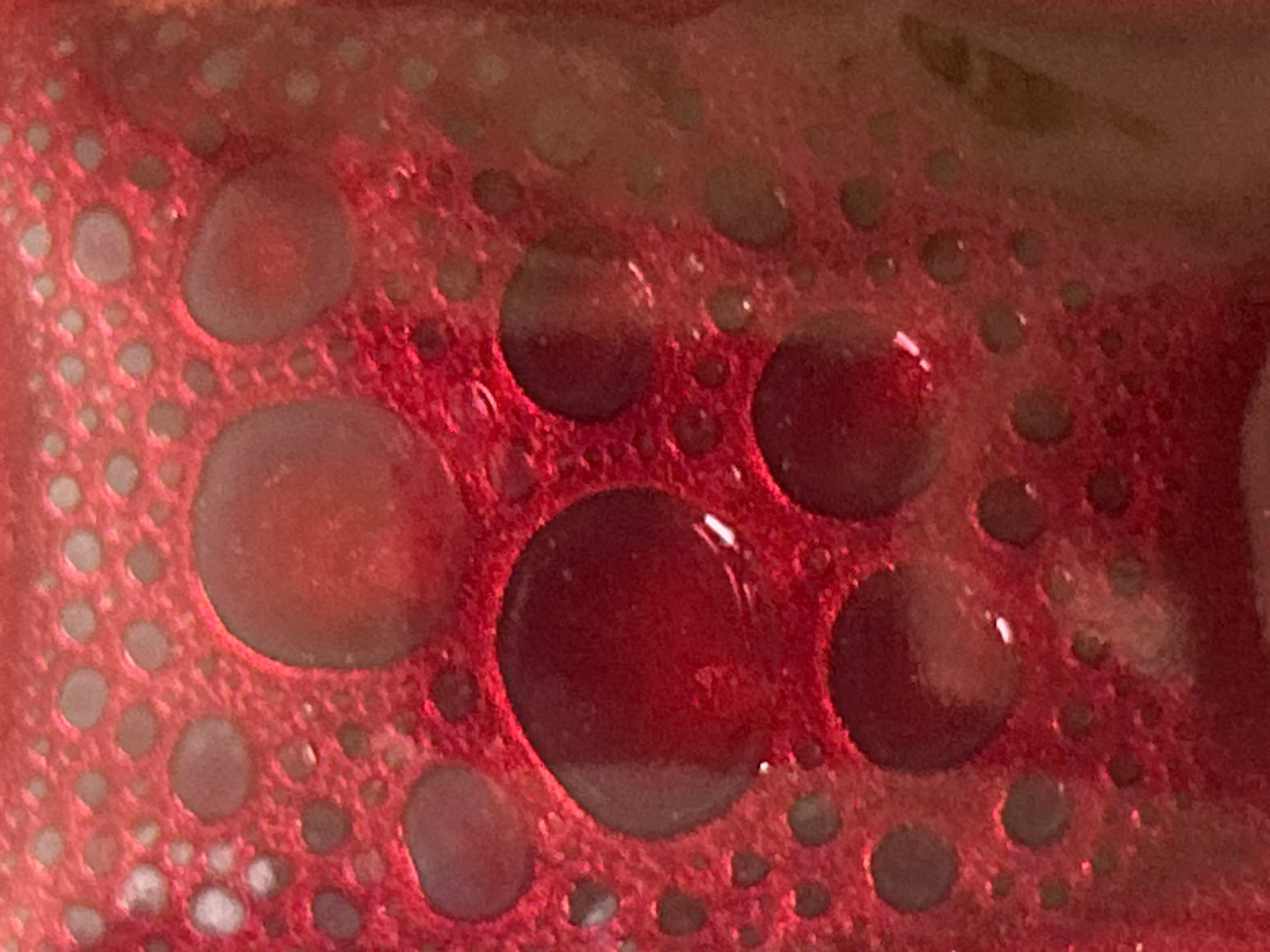 The image shows a close-up of a red, porous material with embedded clear liquid droplets of varying sizes. The material appears to be bubbly or sponge-like, with numerous small holes or pores. Larger droplets are scattered across the surface. There is no indication of the location being in Krumovo, Bulgaria. No people, subjects, objects, actions, or interactions are visible. The environment is limited to the porous, droplet-covered surface.