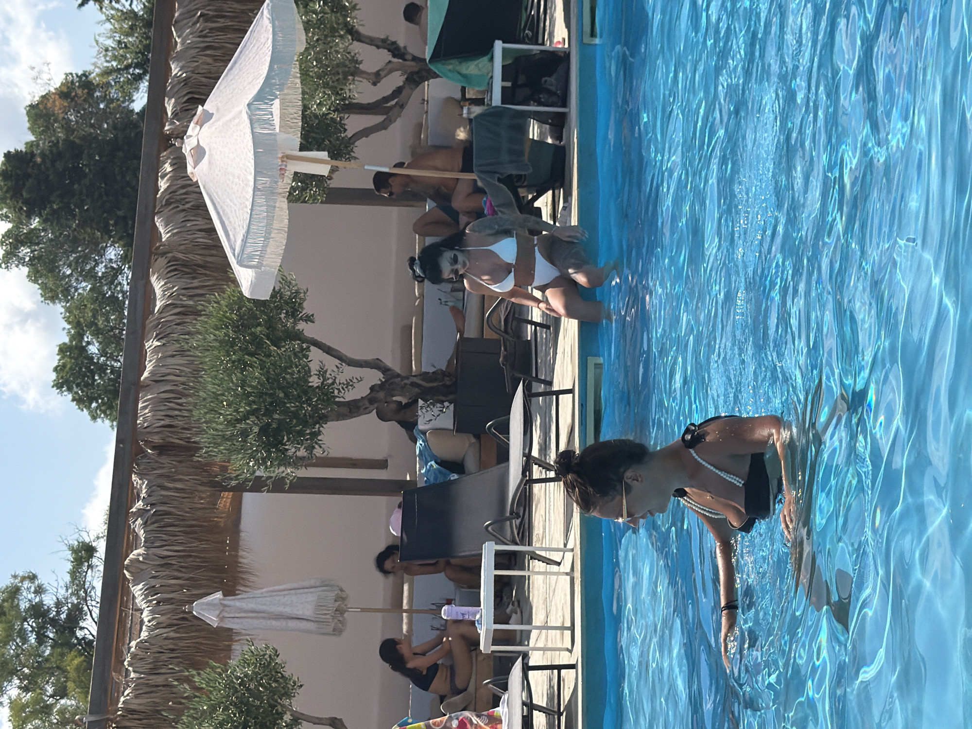 The image depicts a sunny day at an outdoor swimming pool, possibly at a resort or a private villa. In the foreground, a woman with her hair tied up in a bun and wearing sunglasses and a black bikini with pearl straps is in the bright blue water, partially submerged. She is looking to her right and appears to be moving her arms. In the middle ground, another woman with dark hair and sunglasses is sitting on the edge of the pool, dipping her feet into the water. She is wearing a white bikini. Behind her, several other people are relaxing on lounge chairs and a sofa, some under a thatched roof structure. Two olive trees are visible, along with beach umbrellas and lounge chairs scattered around the pool area. The environment suggests a Mediterranean or a similar warm climate setting. The location in Burgas, Bulgaria is not explicitly indicated by any visible landmarks or signs within the image.