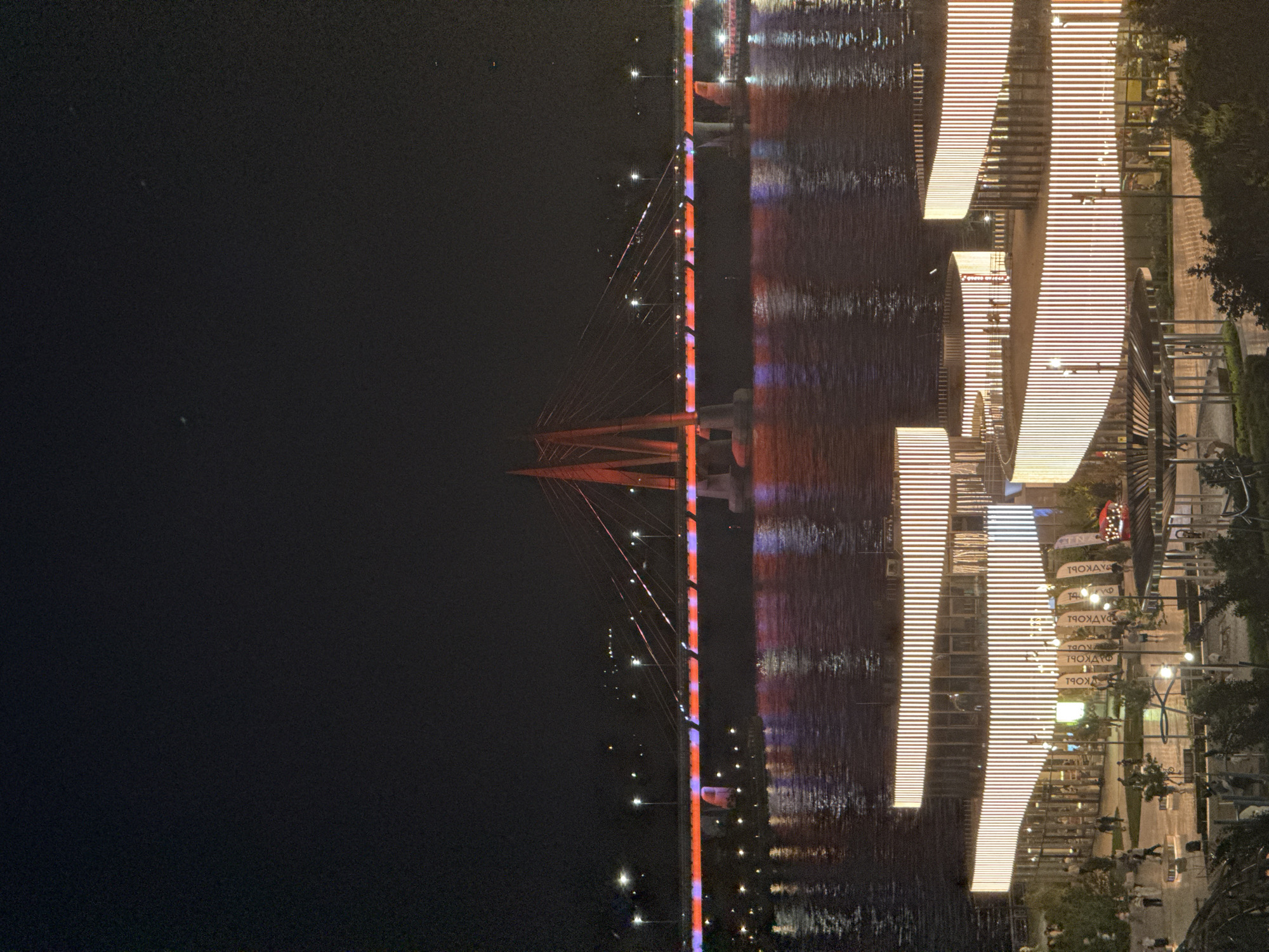 The image shows a nighttime view of a section of the city of Kazan, Russia, featuring the Millennium Bridge and a waterfront area. The bridge is illuminated in red and blue lights, with its reflection visible in the water below. Cables extend from the top of the bridge towards the roadway. The waterfront area features a series of brightly lit, linear structures, possibly buildings or pavilions. The light from these structures is also reflected in the water. People can be seen walking along a paved path adjacent to the structures. To the right of the path, there are trees and greenery. The background is dark, suggesting it is night.