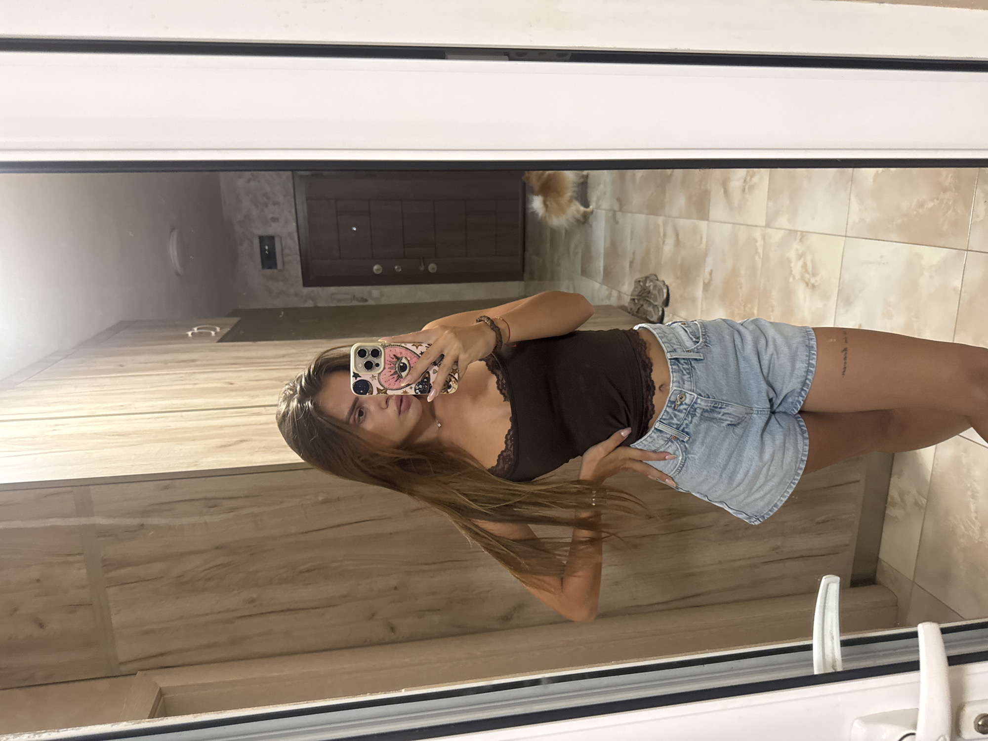 A young woman is taking a mirror selfie in what appears to be an apartment hallway. She has long, brown hair and is wearing a dark brown lace-trimmed camisole and light blue denim shorts. She is posing with her left hand on her hip and her right hand holding her phone, which has a pink case with an eye design. A small, fluffy, light brown dog is visible in the reflection of the mirror in the background, near a closed door. The hallway has light-colored tiled flooring and wooden-paneled walls. The overall impression is that of a casual, domestic setting, possibly in Plovdiv, Bulgaria, given the context provided.