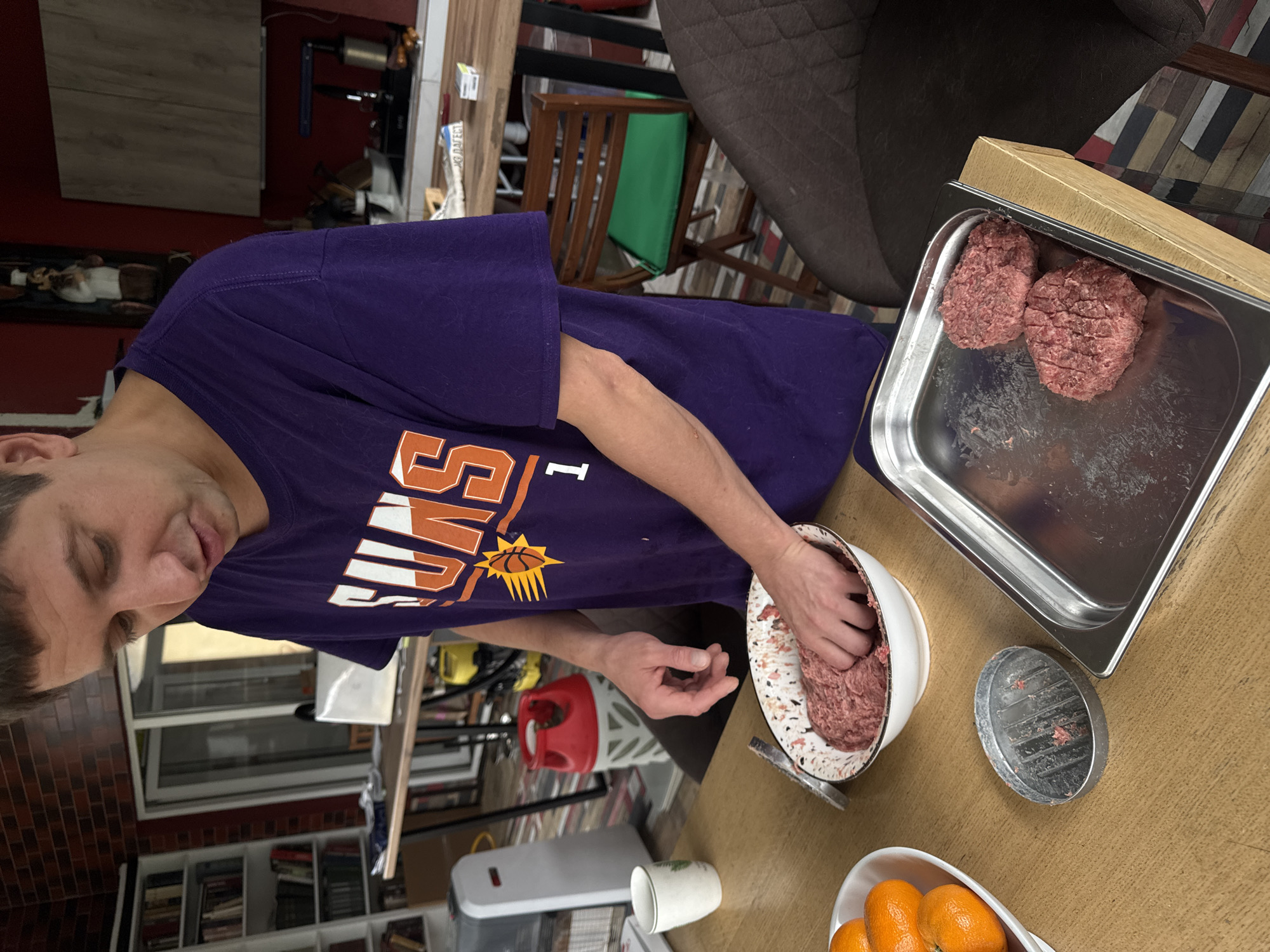 A man wearing a Phoenix Suns T-shirt is making hamburger patties. He is mixing ground beef in a white bowl. To his right is a metal tray holding two hamburger patties. Next to the bowl is a hamburger press and a bowl of oranges. The location is Ptichnoye, Russia.