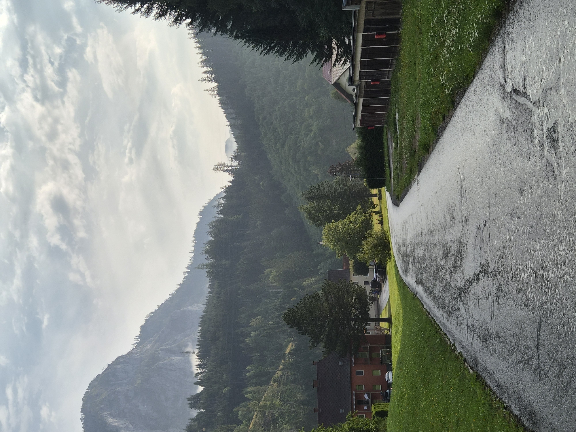 The image shows a wet asphalt road curving through a rural village in Eisenerz, Austria. The road surface is reflecting the overcast sky and appears to have standing water in places. To the left of the road is a green grassy area bordering a red residential building with multiple windows and balconies. Several cars are parked in front of the building. A large evergreen tree stands in front of the building. Further back, a dense forest covers the slopes of a steep, rocky mountain range that dominates the background. The sky is filled with dramatic, cloudy formations. To the right of the road, a row of wooden garages with dark doors is visible, also bordered by grass. The overall impression is a serene, damp, and natural landscape.