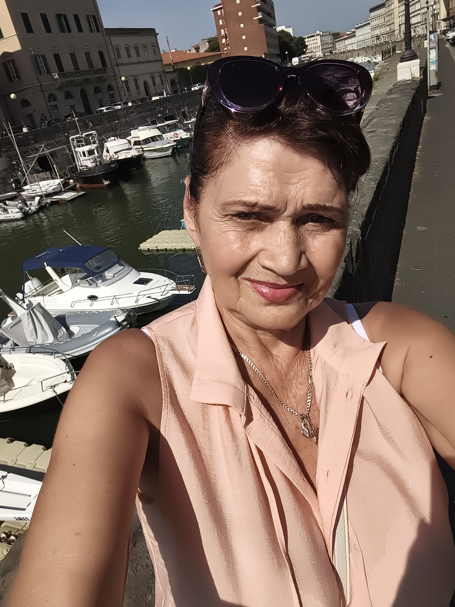 The image shows a woman taking a selfie in a marina in Livorno, Italy. She is wearing a pink blouse and sunglasses. In the background, there are several boats docked, and buildings line the waterfront. The sun is shining, casting shadows on the ground. The marina appears to be in a well-developed urban area, with the architecture suggesting a European city. The water is calm, and the boats are a mix of recreational vessels and what appear to be working boats. The overall impression is one of a sunny day in a scenic Italian port city.