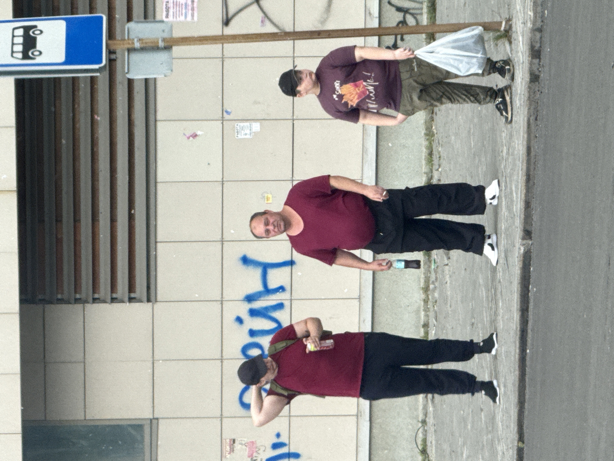 Three individuals are standing on a sidewalk in front of a tiled building wall. To the right, a bus stop sign with a blue background and a white bus icon is visible. The building wall has graffiti in blue spray paint.
The person on the left is a male wearing a dark baseball cap, a maroon t-shirt, a backpack, and black pants. He is holding a clear bottle with a yellow cap in his right hand and has his left hand on the back of his head.
The person in the middle is a male with short hair, wearing a maroon t-shirt and black pants. He is holding a dark bottle in his right hand.
The person on the right is a male wearing a black baseball cap, a dark purple t-shirt with an image of french fries and text, and olive green pants. He is holding a white plastic bag in his right hand and has his left hand in his pocket.
The sidewalk is paved with concrete. The ground in front of the sidewalk is asphalt. The environment appears to be an urban setting.