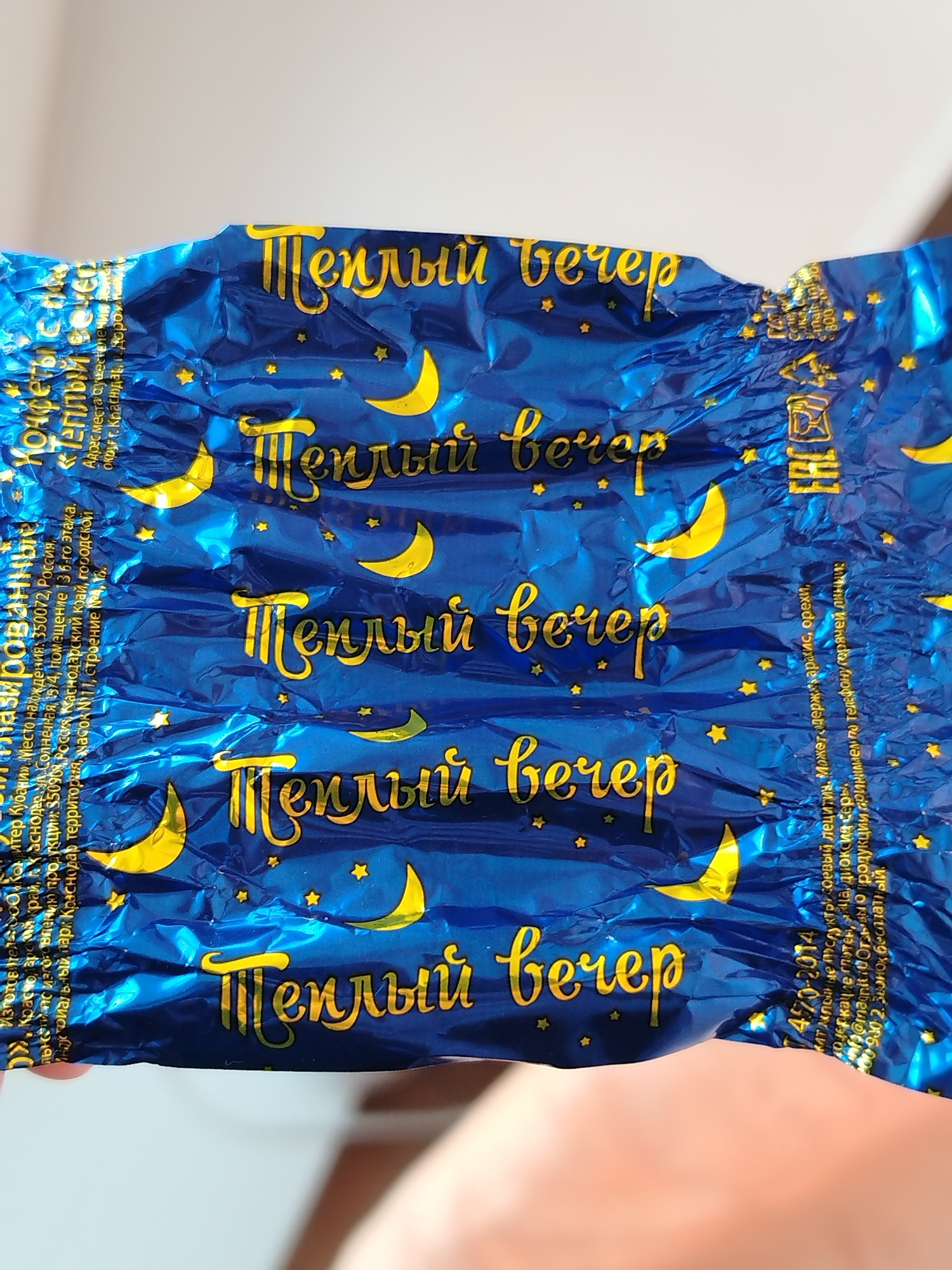 The image shows a candy wrapper with the words "Теплый вечер" (Warm Evening) written multiple times in gold lettering on a blue background decorated with crescent moons and stars. Information printed on the wrapper in Cyrillic alphabet includes the manufacturer information as "ЗАО Тандер. "Магнит" (Magnit) and address in Краснодар (Krasnodar), Russia.