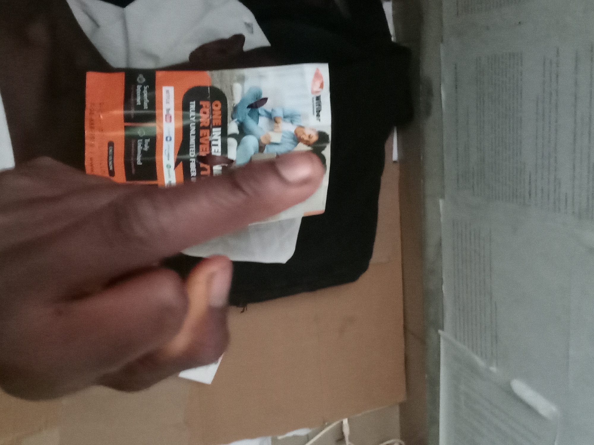 A close-up shot shows a dark-skinned human hand holding a promotional card for Witribe internet services. The card features an image of a young person using a laptop. The surrounding environment includes clothing items and cardboard. There is insufficient information to determine the location within Akwete, Nigeria.