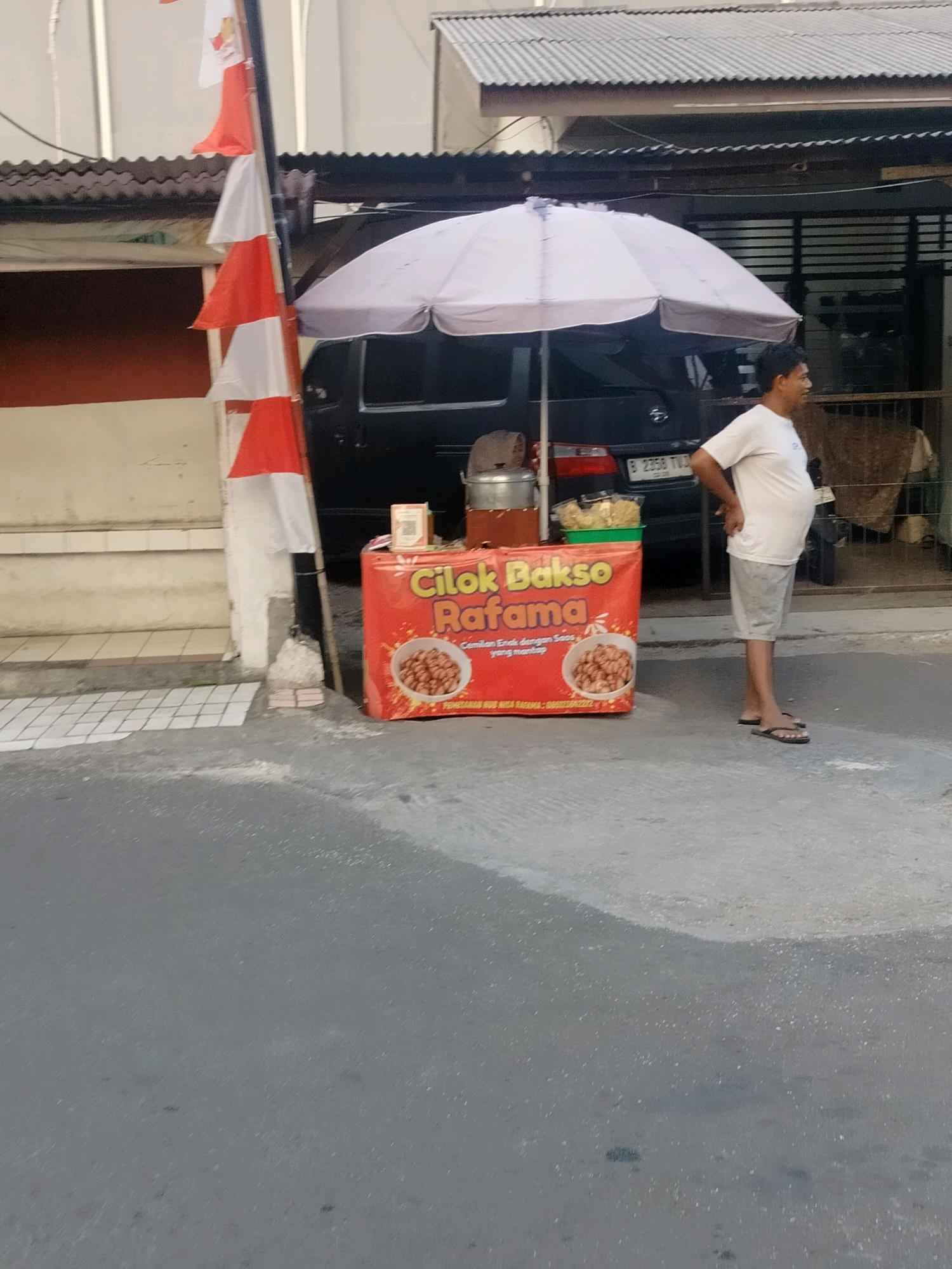 An Indonesian man stands next to a red food cart selling "Cilok Bakso Rafama" on a street in East Jakarta, Indonesia. The cart is adorned with an image of "cilok" (a type of Indonesian meatballs) and text that reads "Cemilan Enak dengan Saus yang mantap" (Delicious snacks with delicious sauce). A white umbrella is propped over the cart, shading it. Behind the cart, a black car is parked, and a national flag of Indonesia is displayed on a flagpole. The ground is paved with concrete, and a building with a corrugated metal roof is visible in the background. The man is wearing a white t-shirt and light grey shorts, and he is looking to his right.